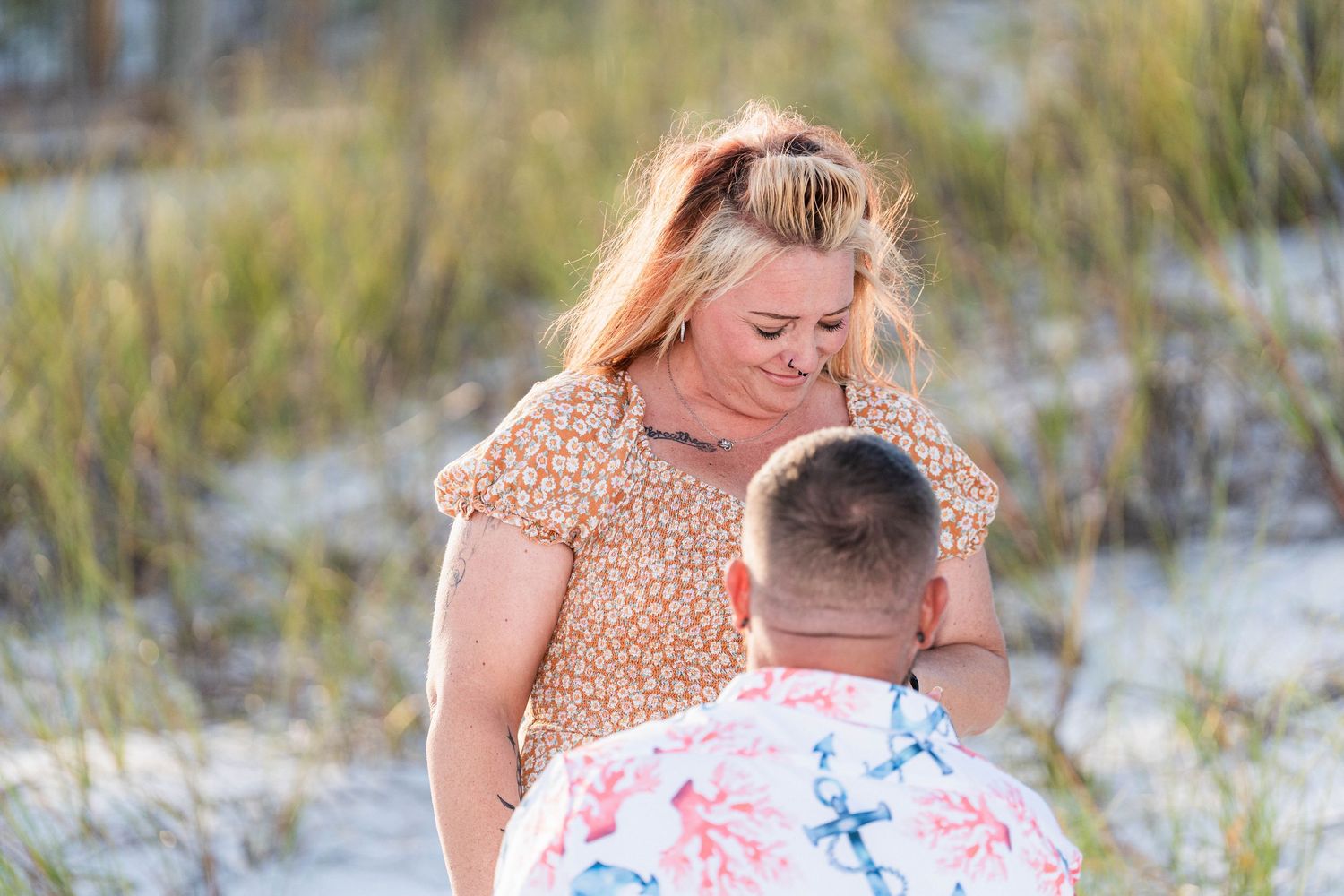 Couple sharing a romantic moment on a sandy beach with sea grass, woman smiling down at man in floral shirt.