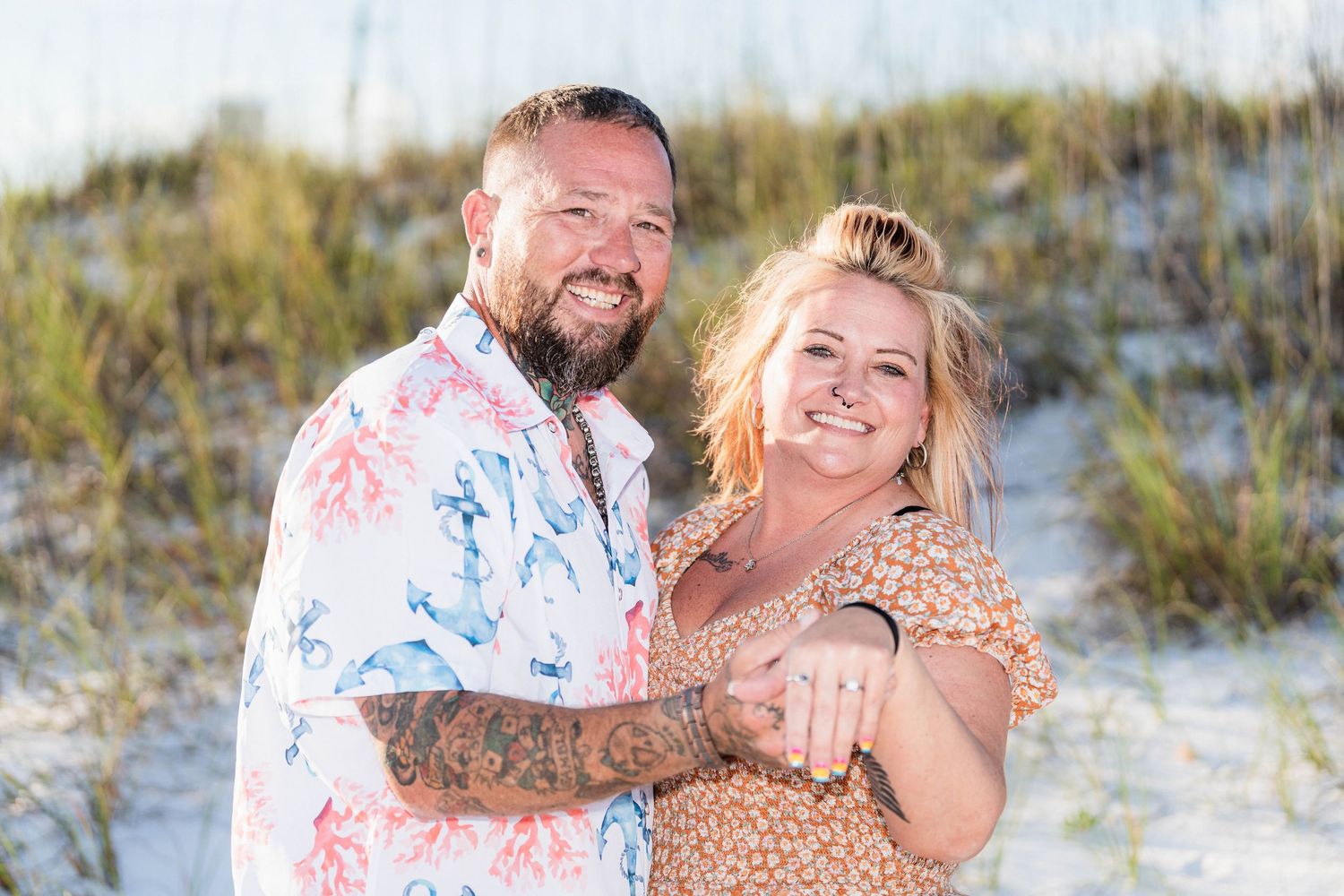 Happy couple embracing on a sandy beach with dunes and sea grass, man in floral shirt with tattoos, woman in floral dress.