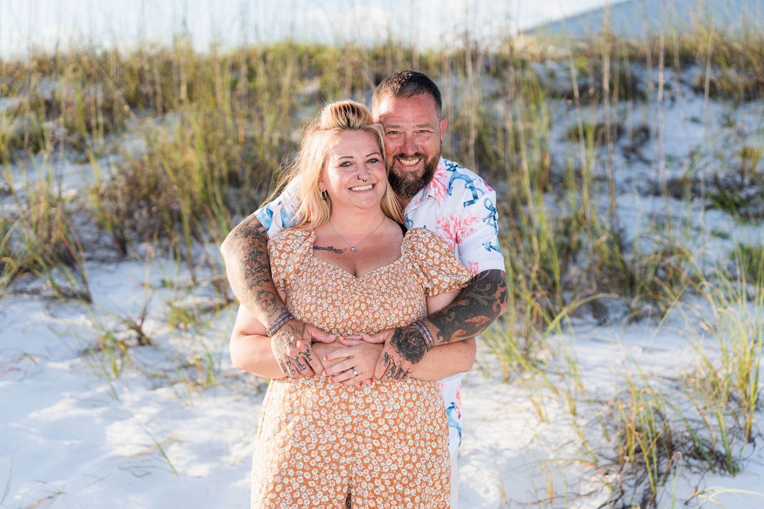 Happy couple embracing on white sand beach dunes, woman in floral romper and tattooed man in colorful shirt.