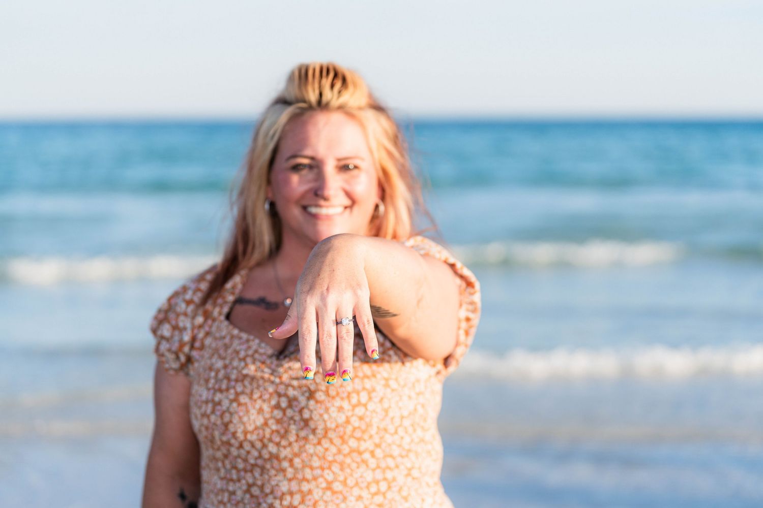 Smiling blonde woman at beach showing engagement ring, wearing floral dress with ocean waves in background.