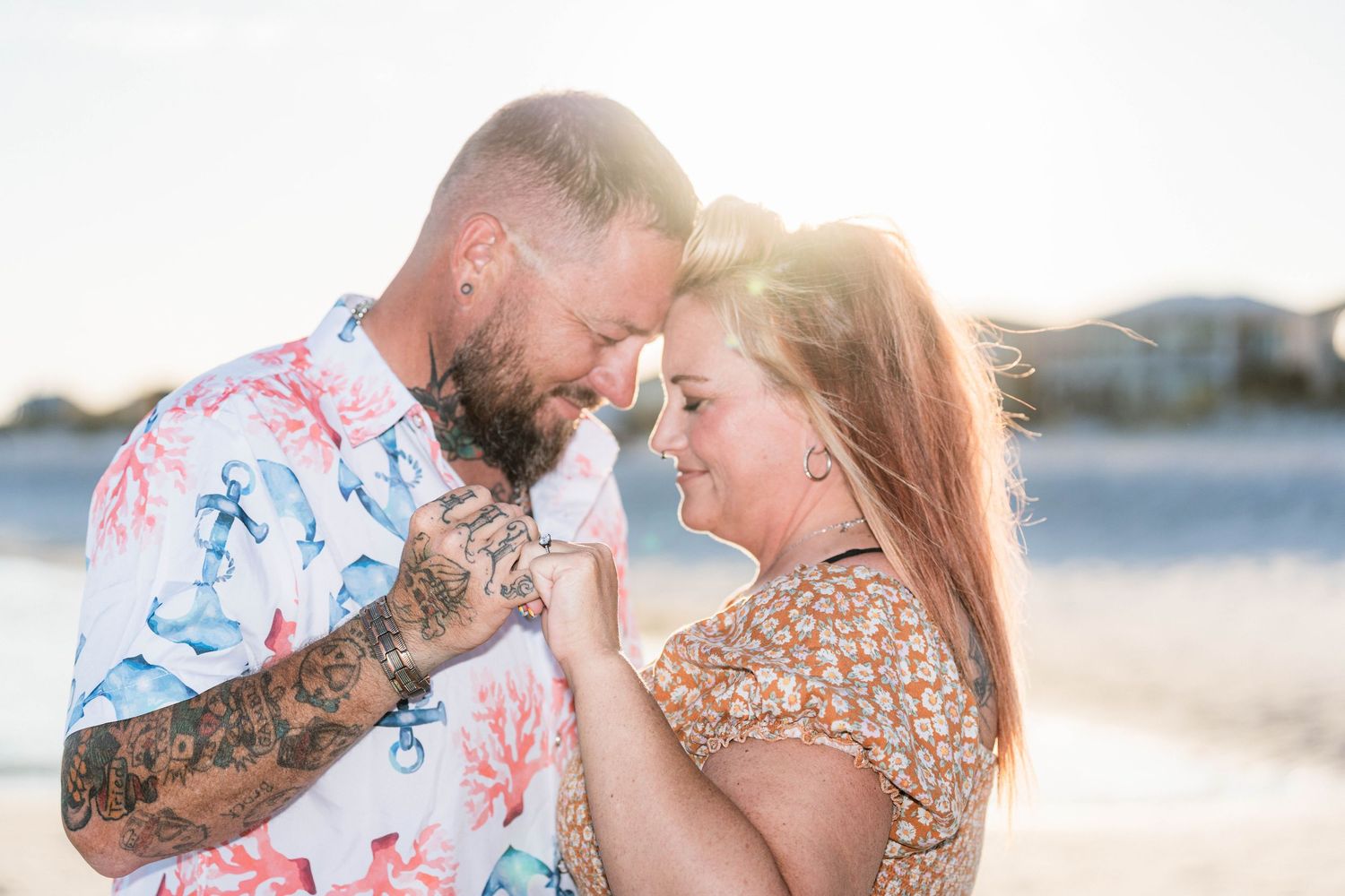 Couple sharing a romantic forehead touch on a sunny beach, man with tattoos in floral shirt, woman in patterned dress.
