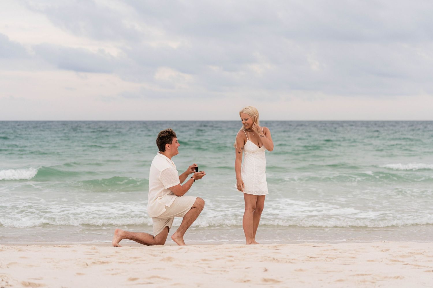 Man proposing to woman on a sandy beach with turquoise ocean waves in the background.