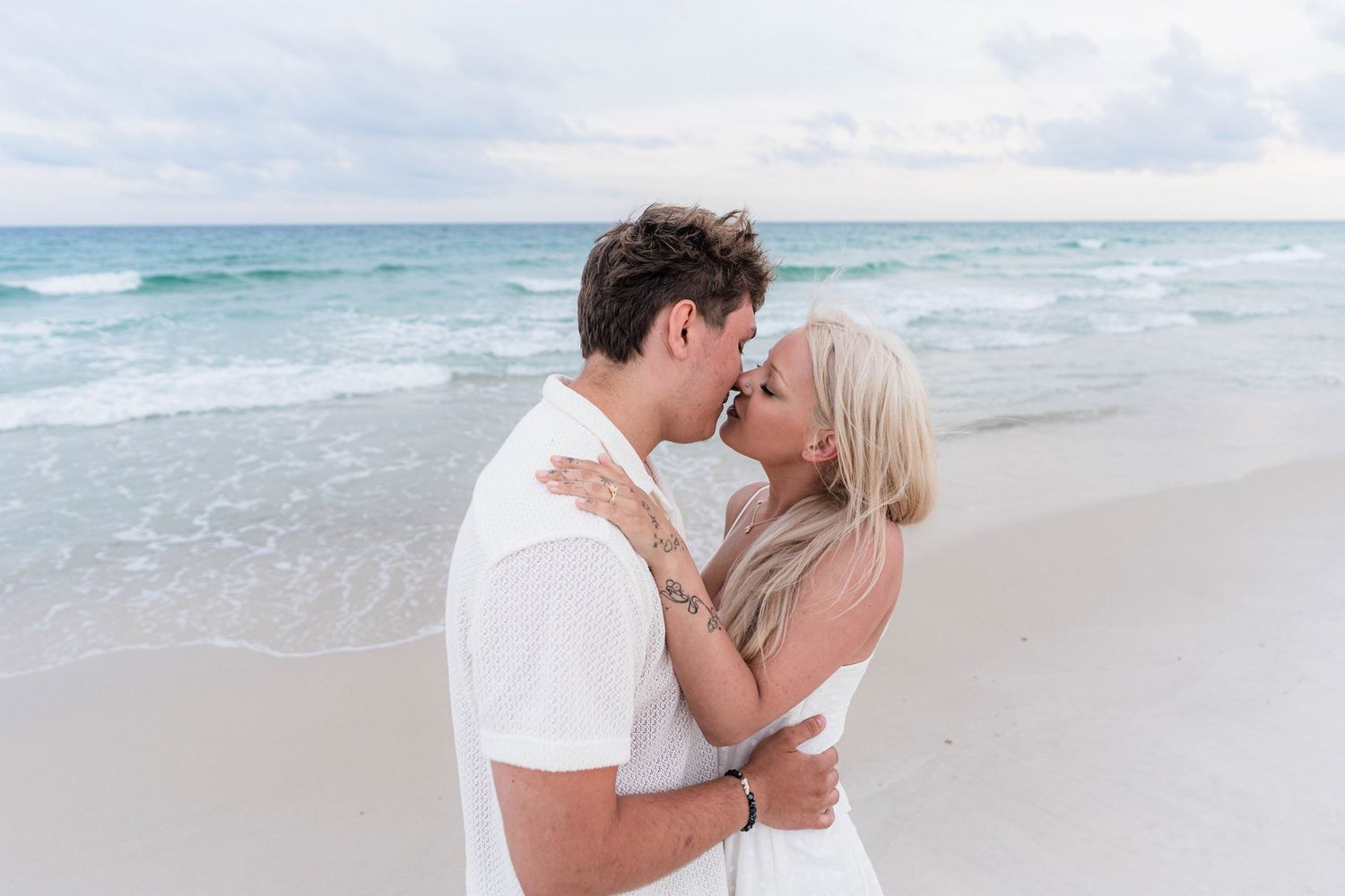 Couple sharing a romantic kiss on a white sand beach with turquoise ocean waves in the background.