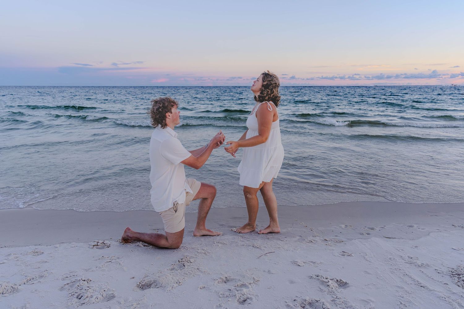 A couple in white attire shares a romantic beach proposal at sunset, with gentle ocean waves in the background.