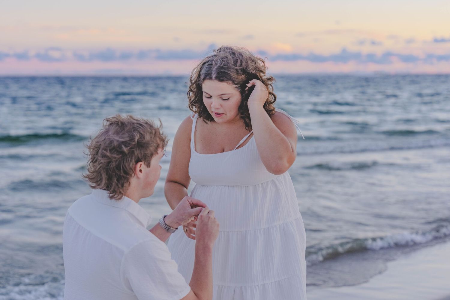 Couple in white outfits sharing a romantic beach proposal at sunset with ocean waves in the background.