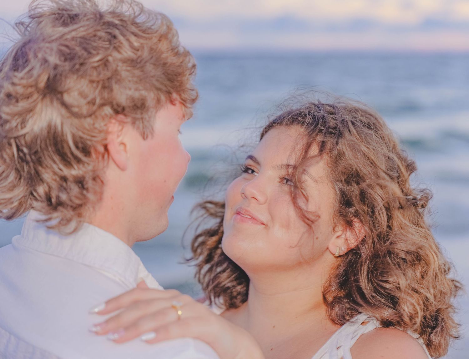 Romantic couple embracing on a beach at sunset, both with curly hair wearing white, smiling at each other.