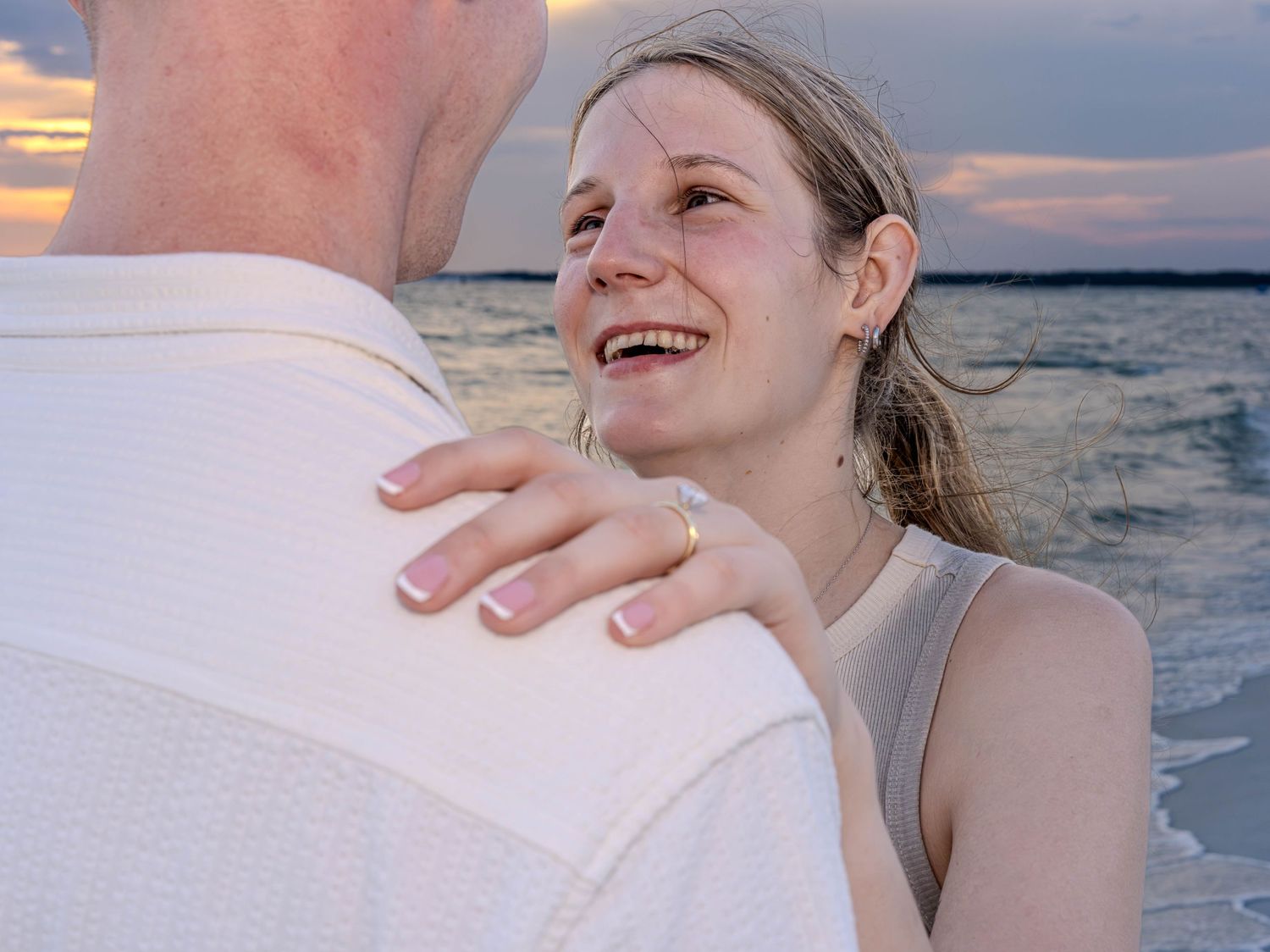 Couple embracing at sunset on the beach, woman smiling and showing engagement ring on her hand.