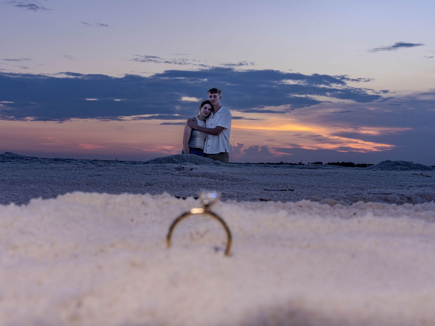 Couple embracing on a beach at sunset with an engagement ring in the foreground on white sand.
