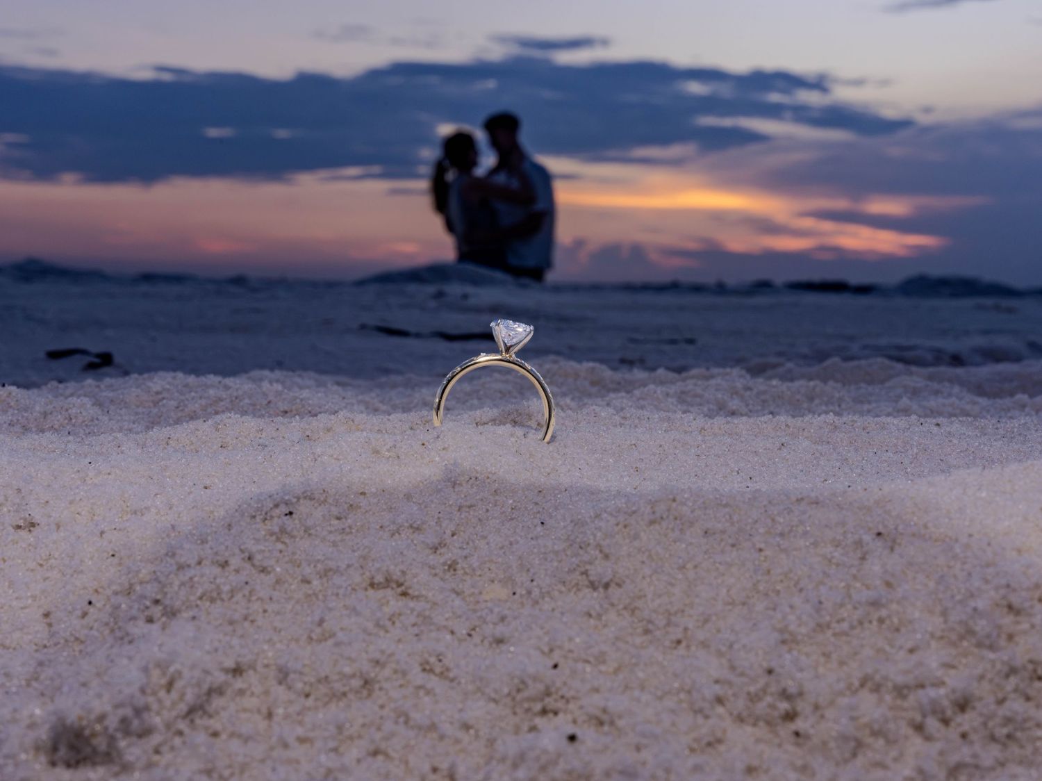 Diamond engagement ring in beach sand at sunset with couple embracing in soft focus background.