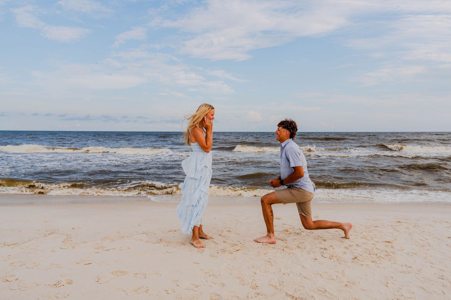 Man kneeling on one knee proposing to woman in white dress on a sandy beach with ocean waves in the background.