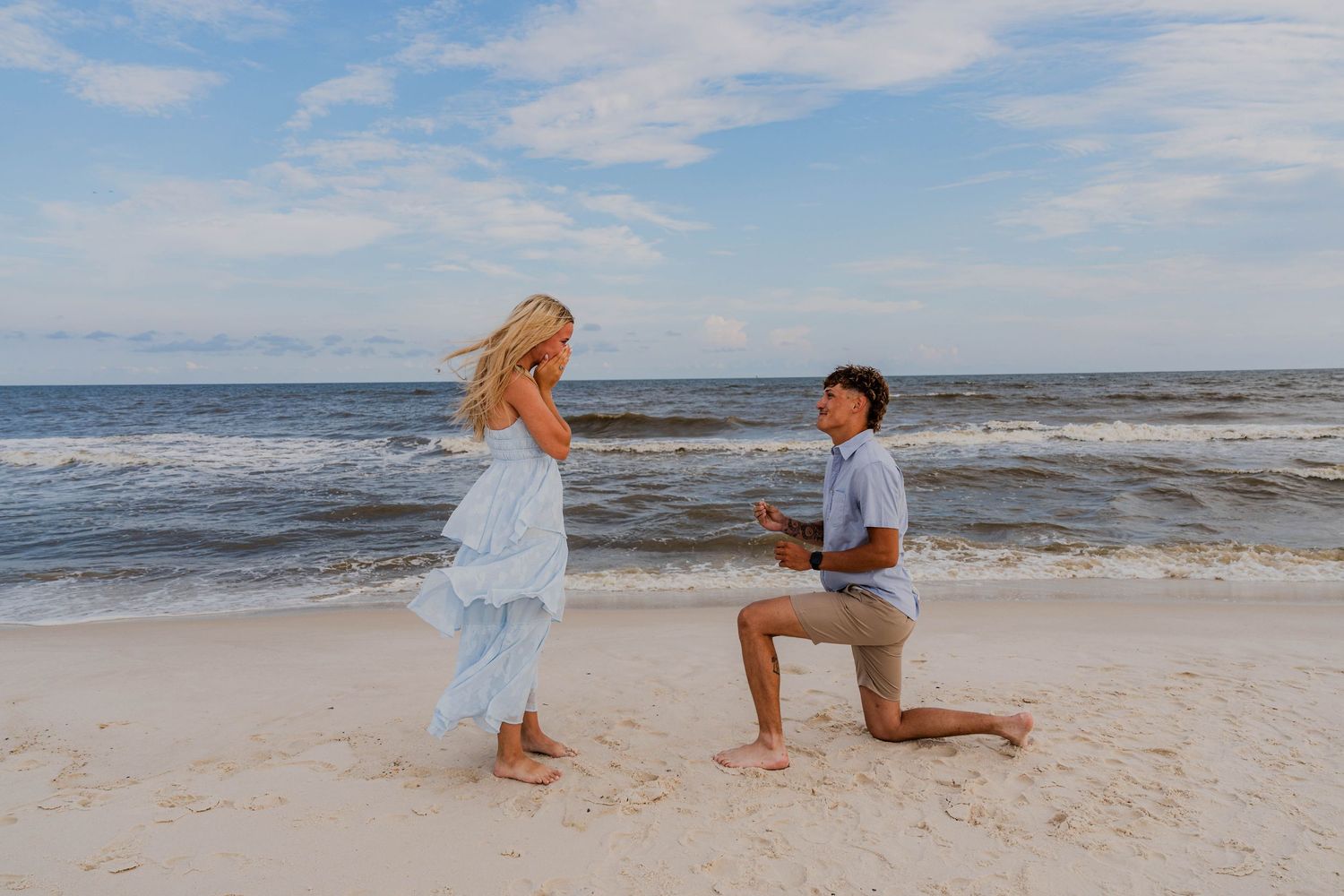 Man proposes on bended knee to woman in blue dress on sandy beach with ocean waves in background.