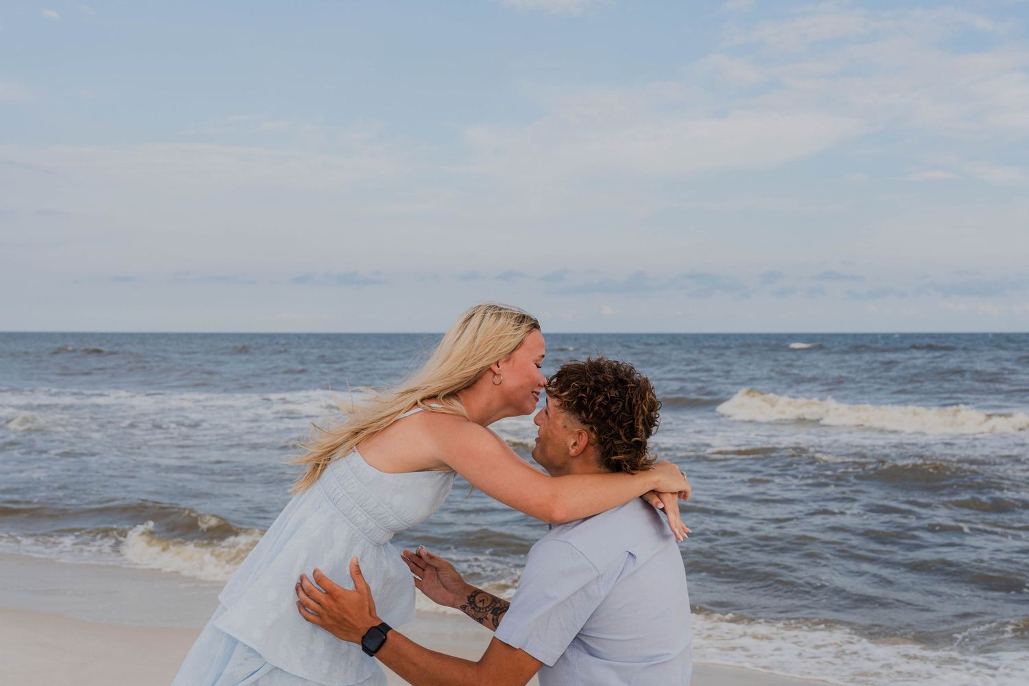Couple sharing a romantic kiss on a sandy beach with ocean waves in the background on a cloudy day.