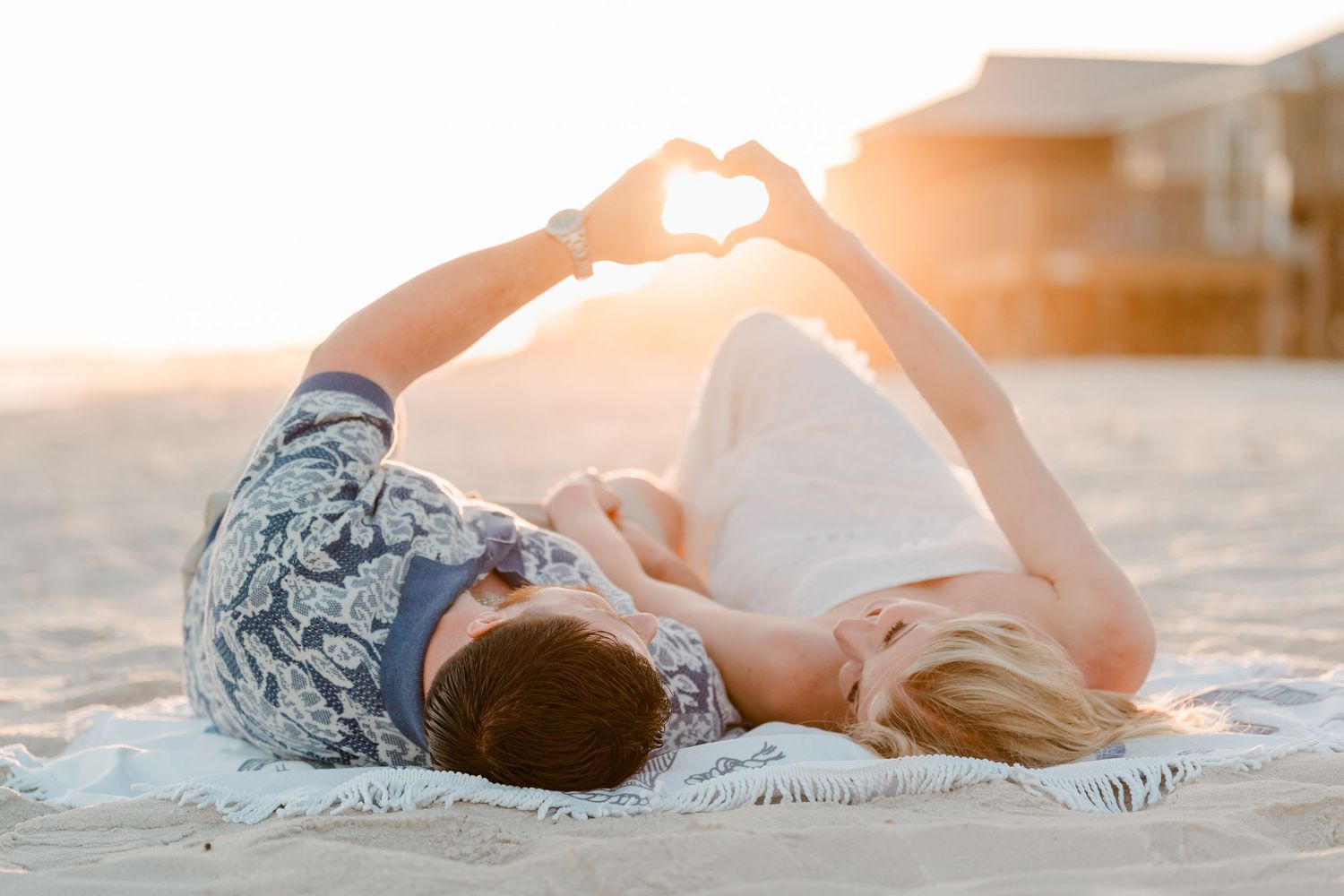 Couple lying on beach blanket making heart shape with hands at golden sunset.