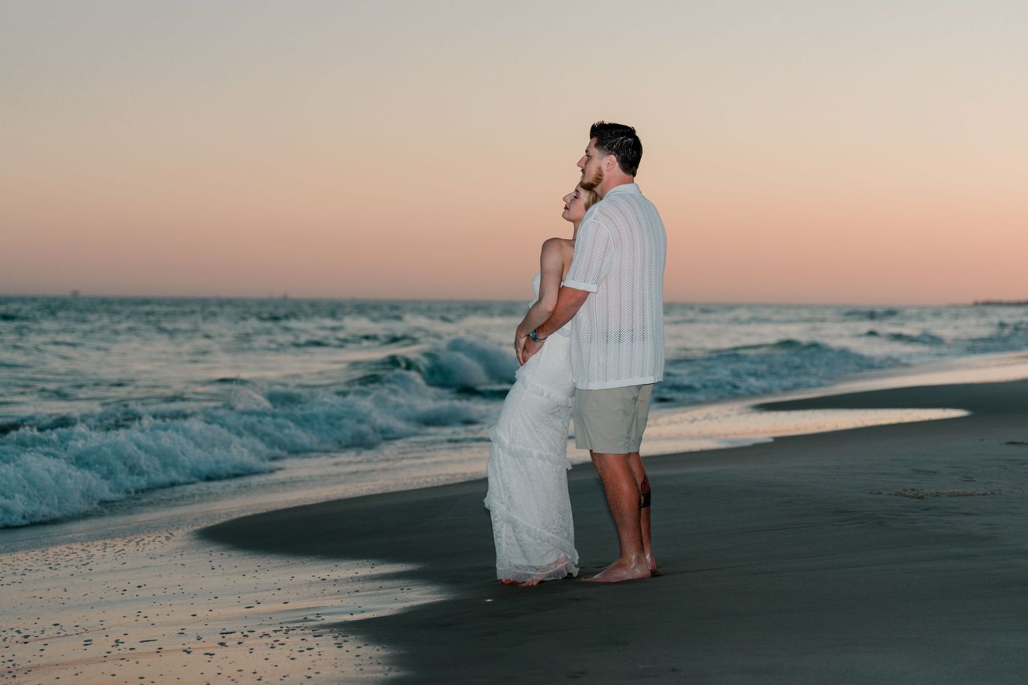Couple embracing on a sandy beach at sunset with gentle ocean waves in the background.