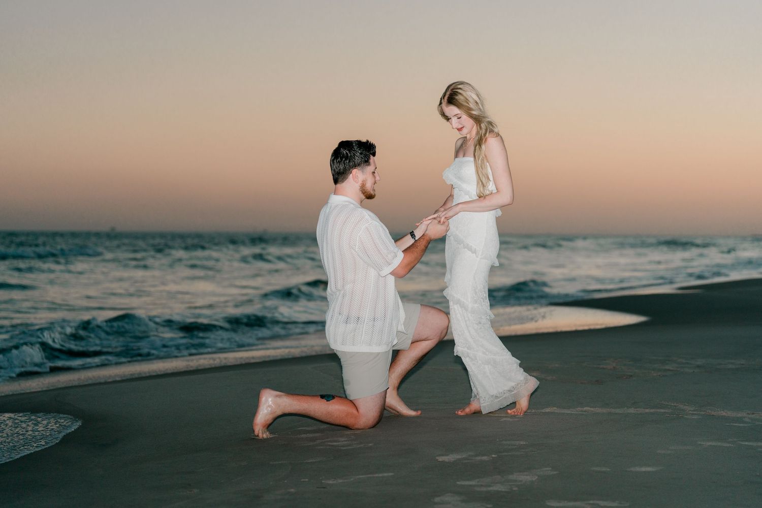 Man proposing to woman on beach at sunset, both dressed in white, romantic oceanside marriage proposal scene.