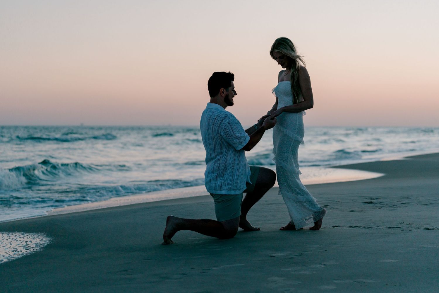 Man kneeling on beach proposing to woman at sunset, romantic ocean backdrop with soft pink sky.