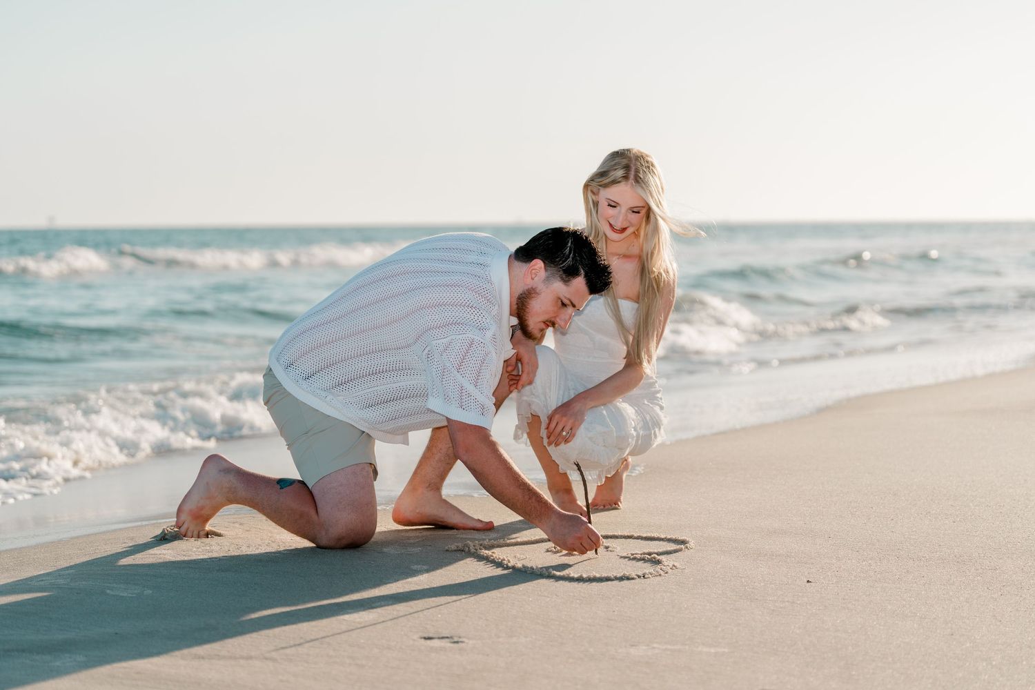 Couple drawing a heart in the sand on a sunny beach, smiling and enjoying a romantic moment by the ocean.