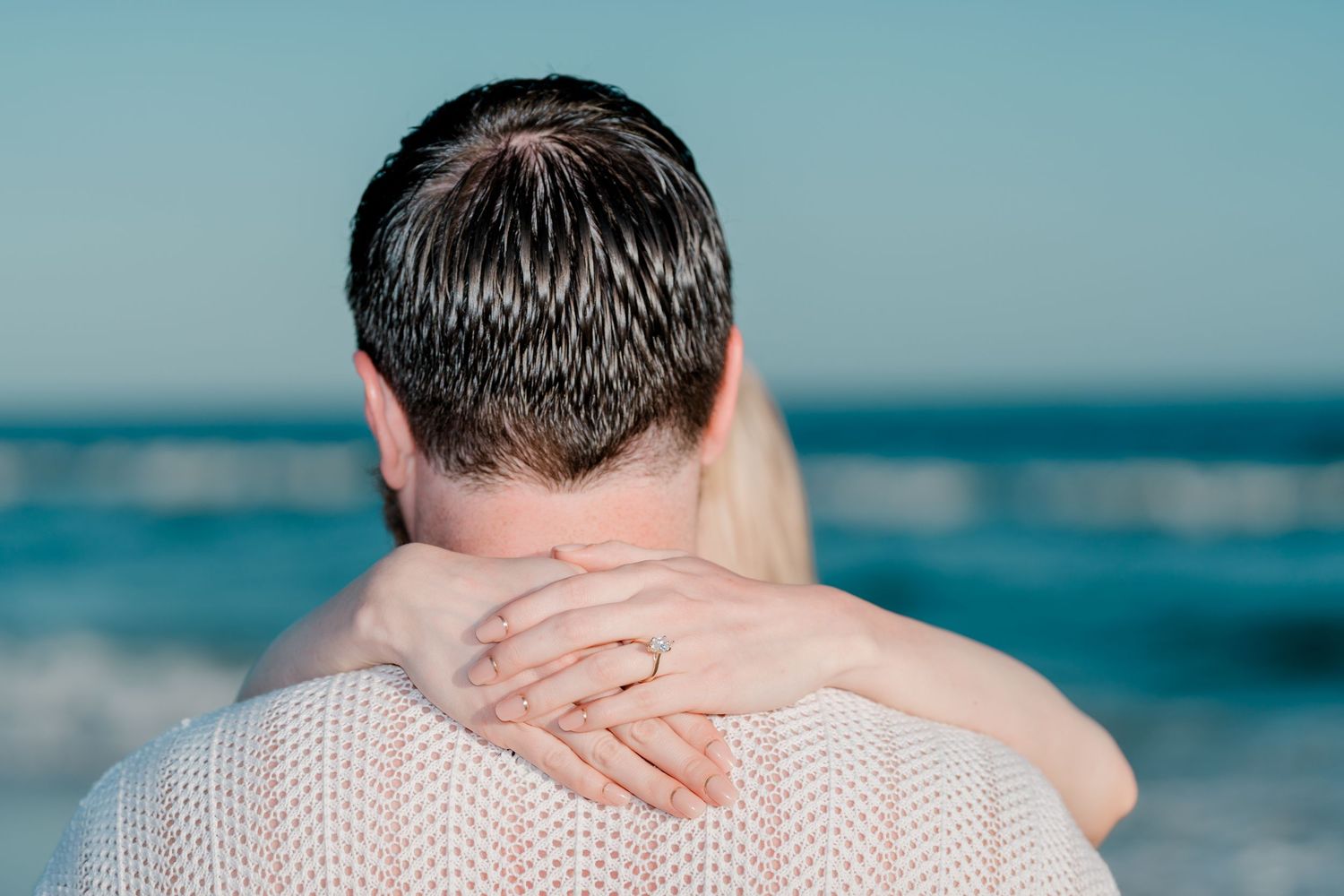 Couple embracing at the beach, woman wearing a gold ring, ocean waves in the background on a clear day.