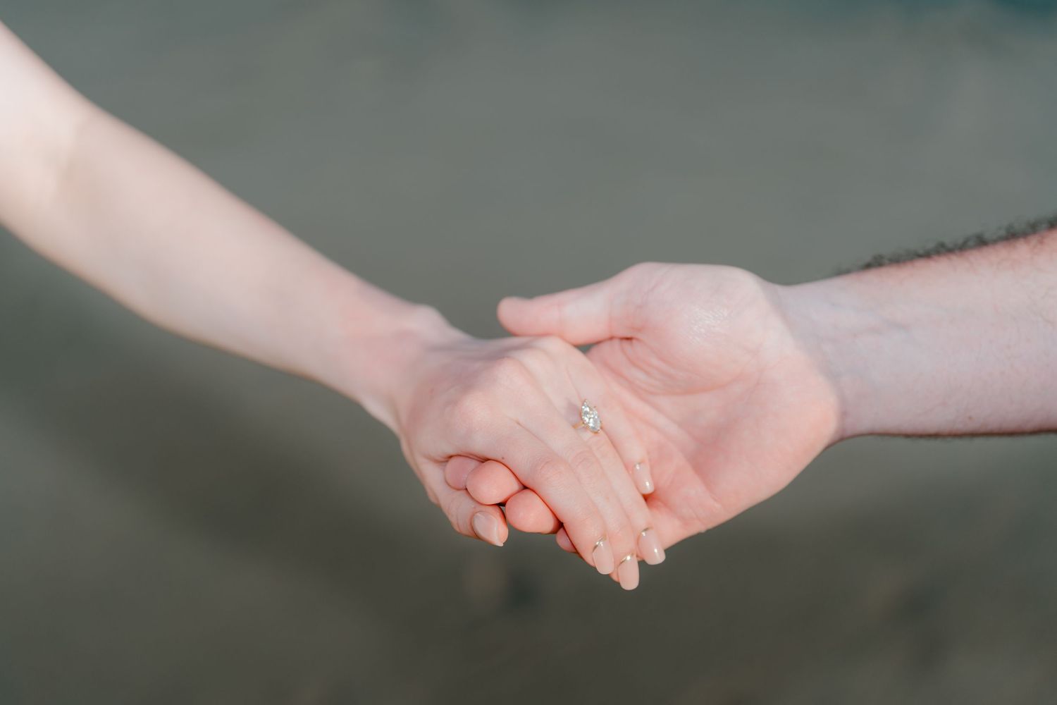 Close-up of a couple holding hands, woman wearing a diamond engagement ring, soft blurred background.