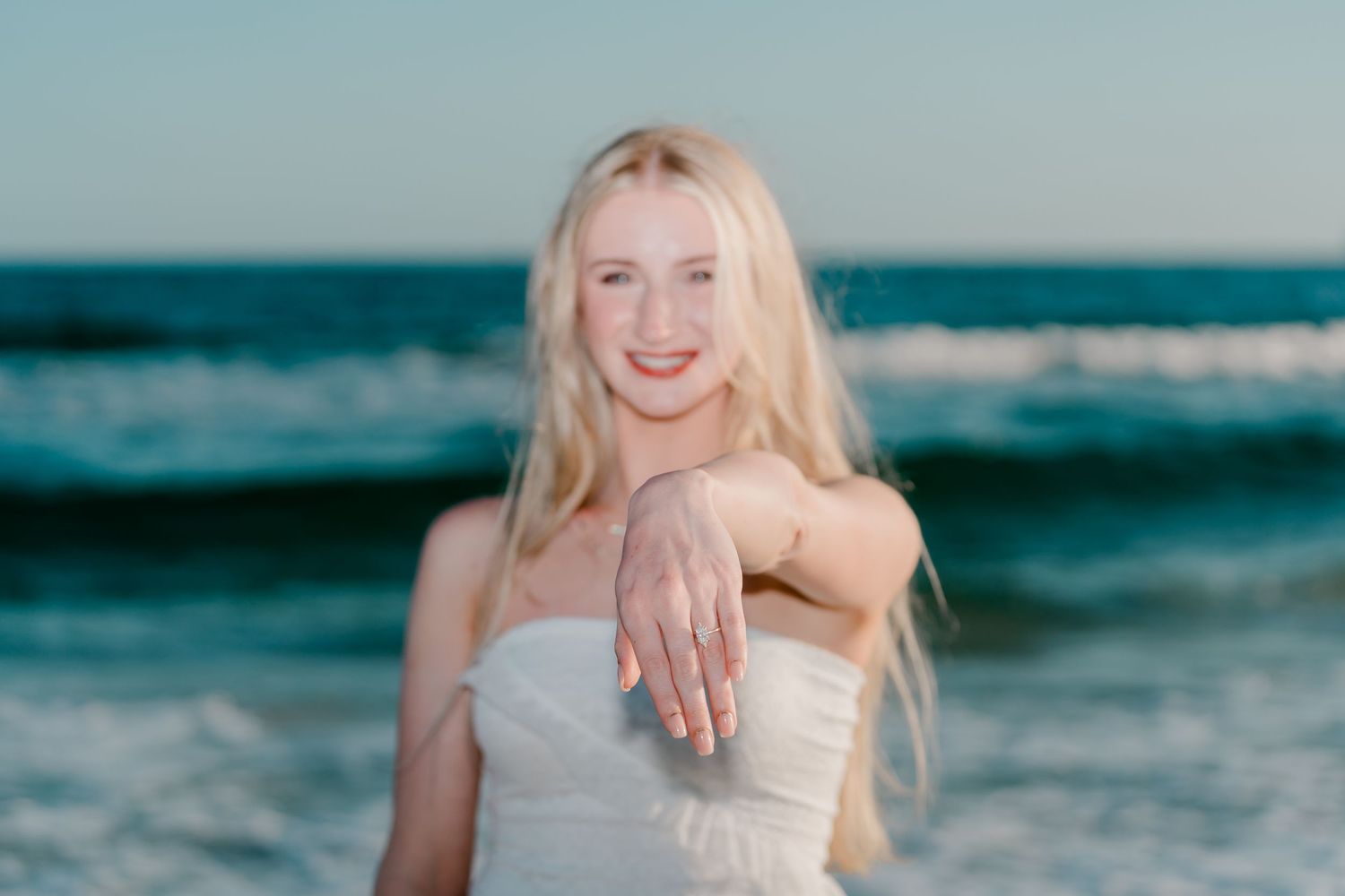 Blonde woman in white dress showing engagement ring on beach with ocean waves in background.