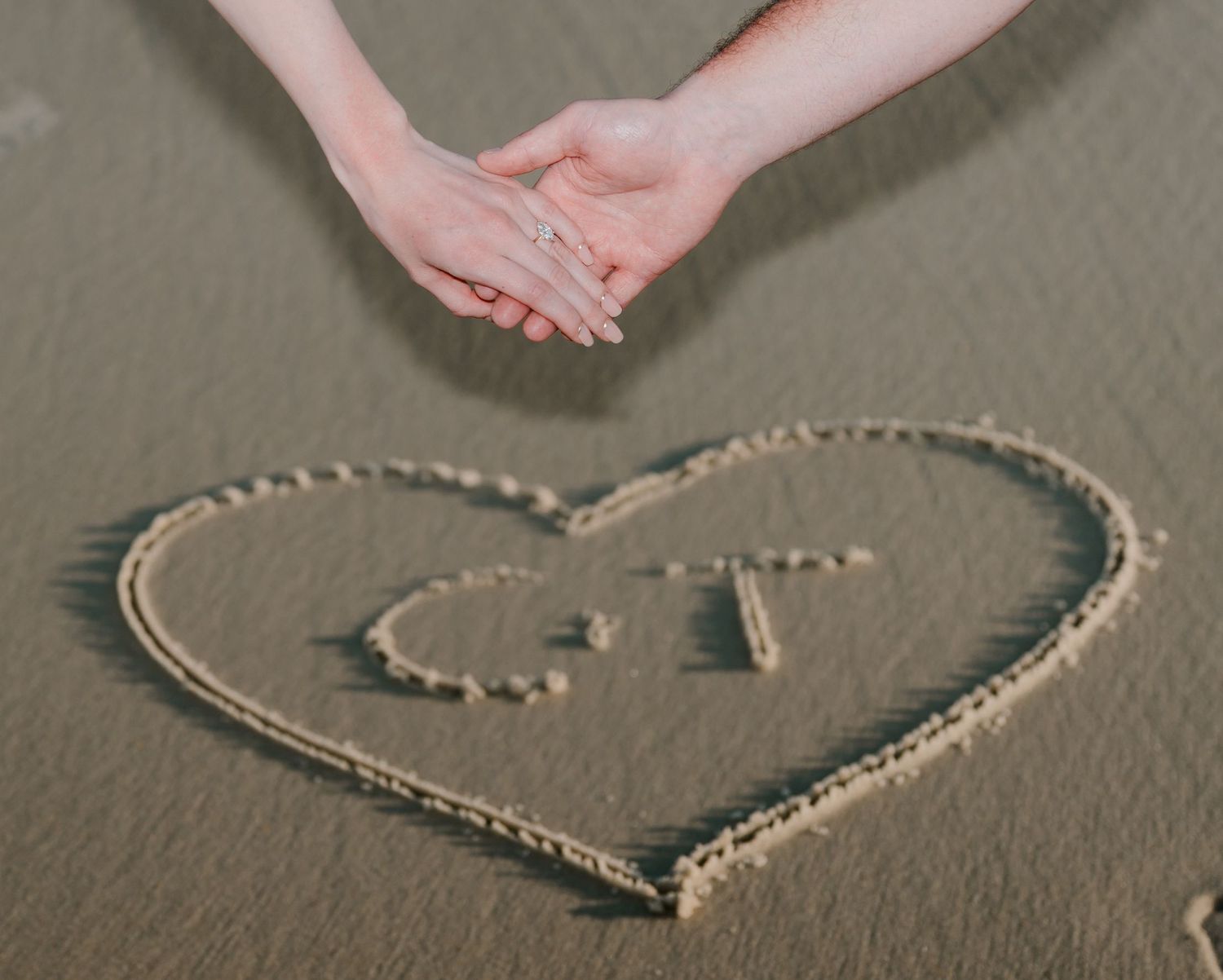 Couple holding hands over a heart drawn in sand with initials C and T at a romantic beach setting.