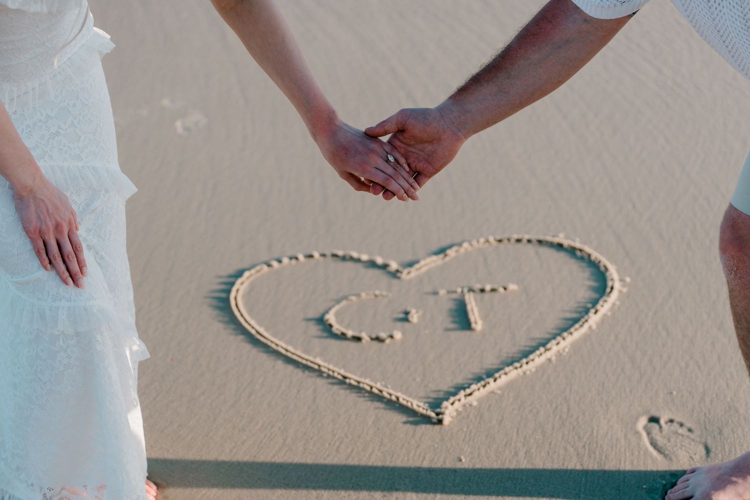 Couple holding hands on beach with heart drawn in sand containing initials C and T.