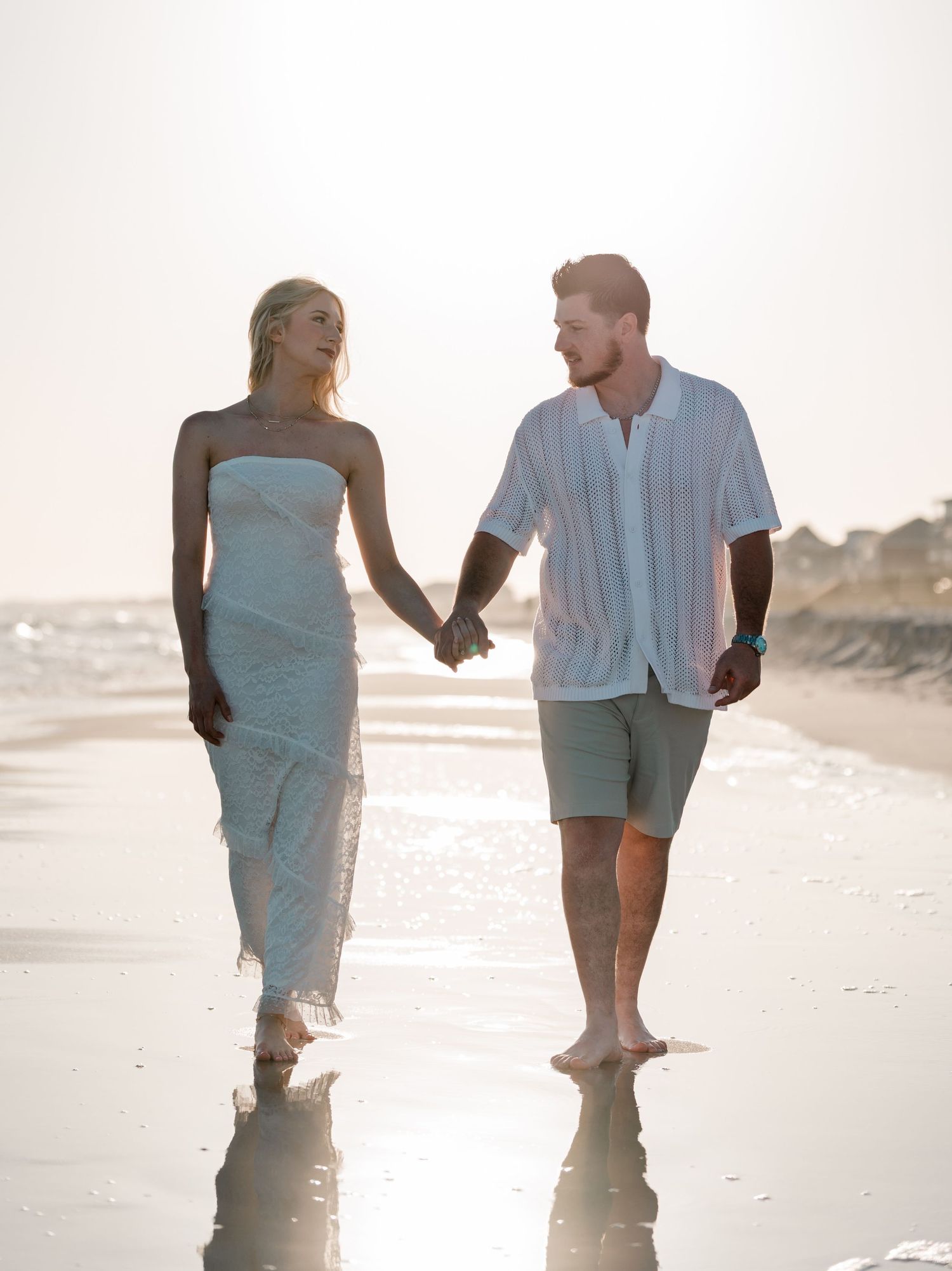 Couple holding hands walking barefoot on a sunny beach, woman in white dress, man in white shirt and shorts.