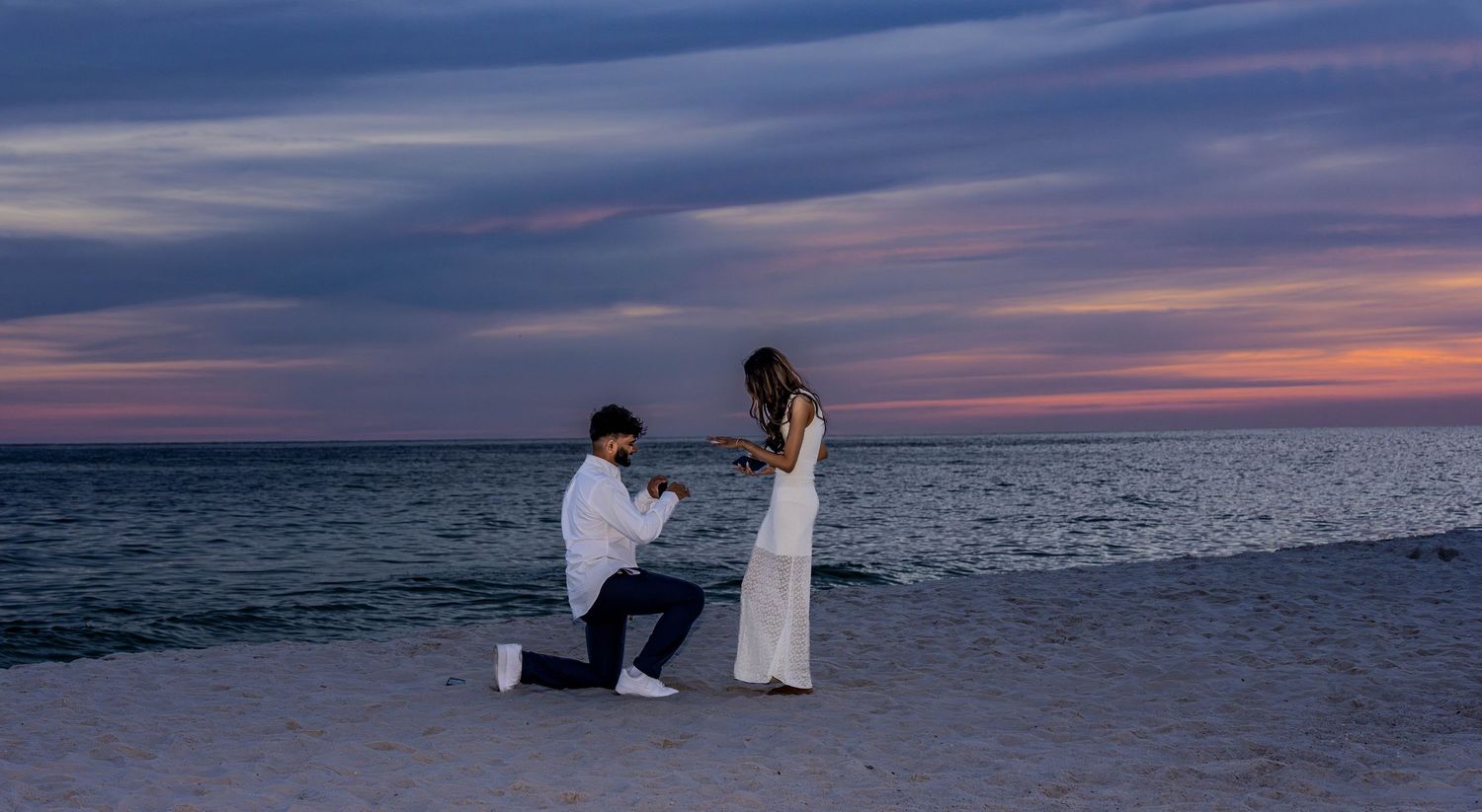 Man proposes to woman on a romantic beach at sunset, kneeling in the sand with colorful sky backdrop.