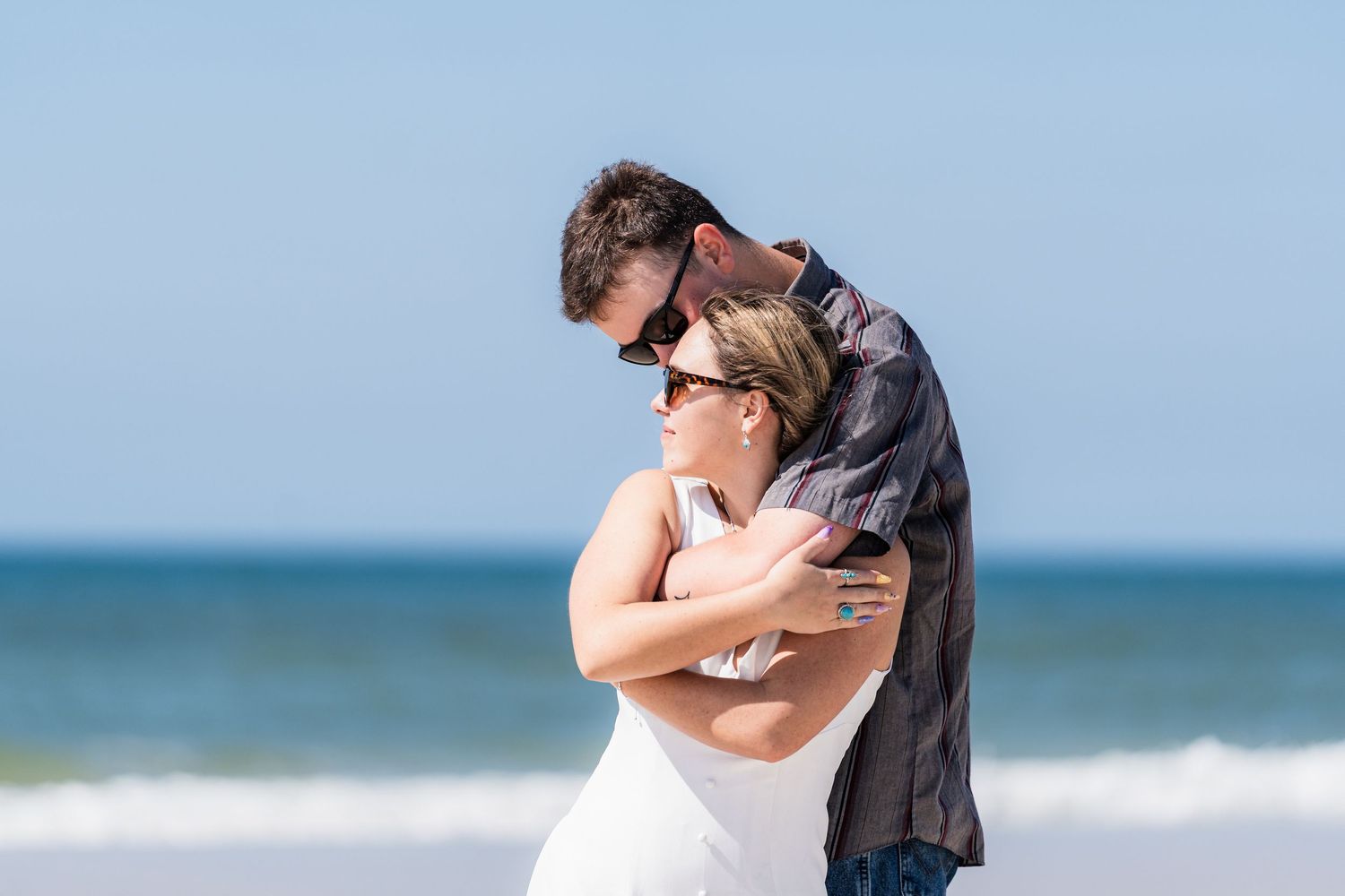 Couple embracing romantically on a sunny beach with ocean waves in the background, both wearing sunglasses.