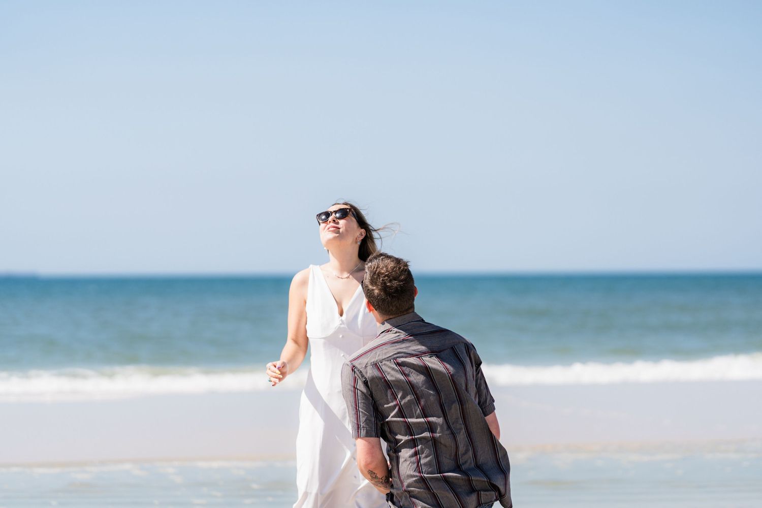 Couple sharing a romantic moment on a sunny beach, woman in white dress laughing joyfully with ocean waves behind them.