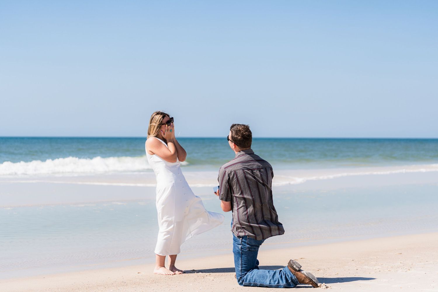 Man proposes to woman on a sunny beach, kneeling in the sand with ocean waves in the background.