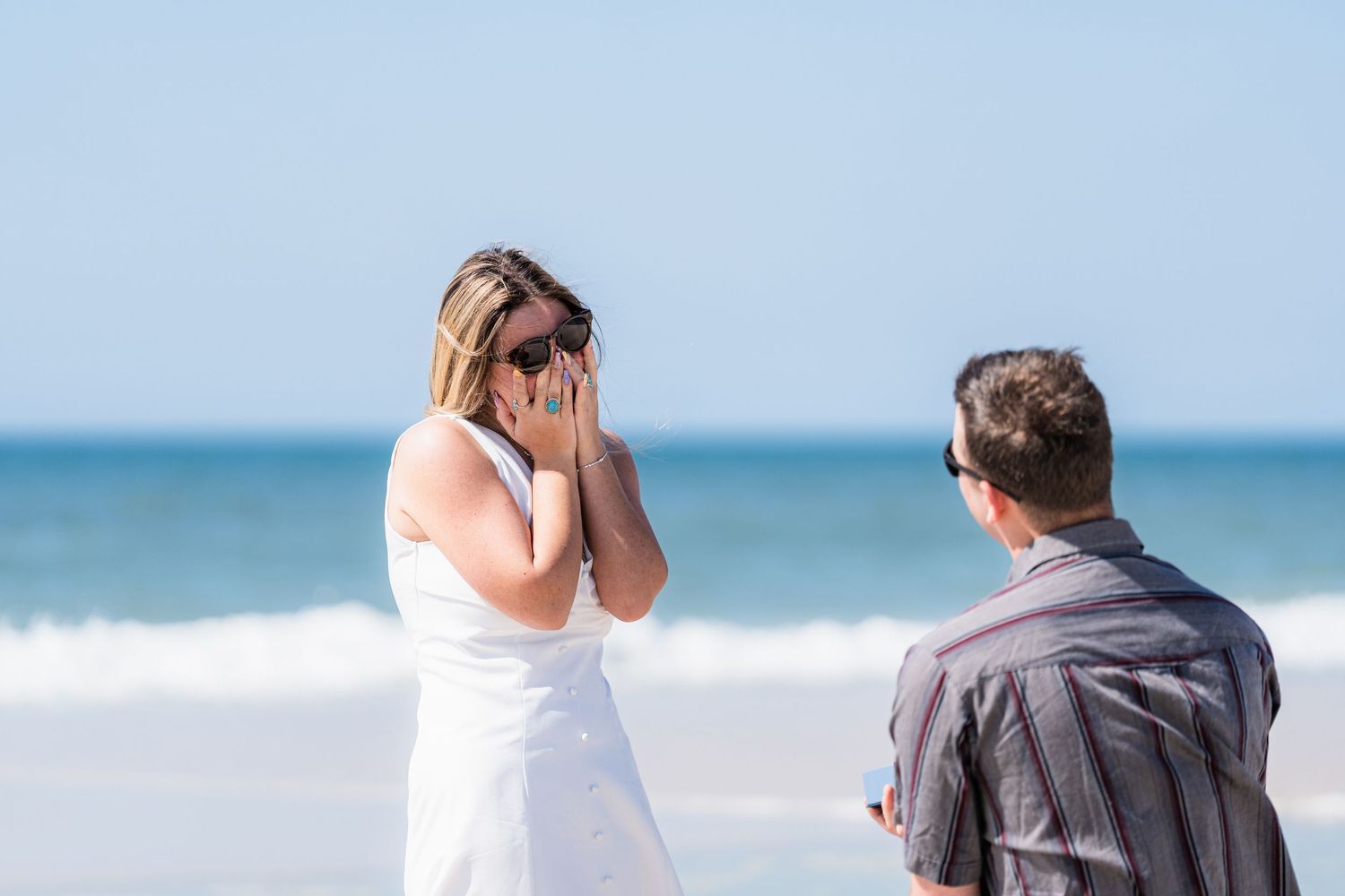 Woman in white dress crying tears of joy during a beach marriage proposal on a sunny day.