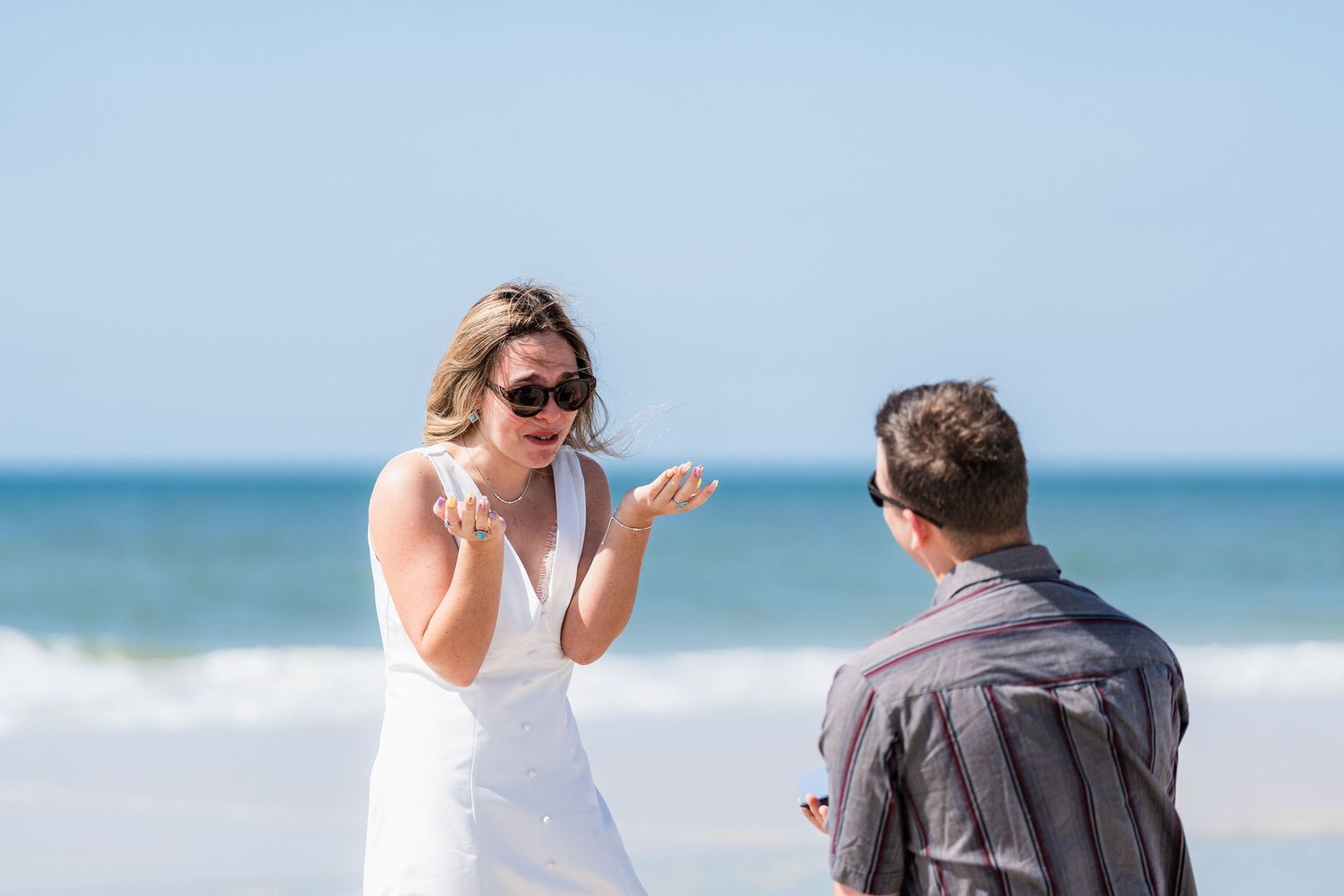 Surprised woman in white dress reacts to beach marriage proposal with hands clasped near ocean.
