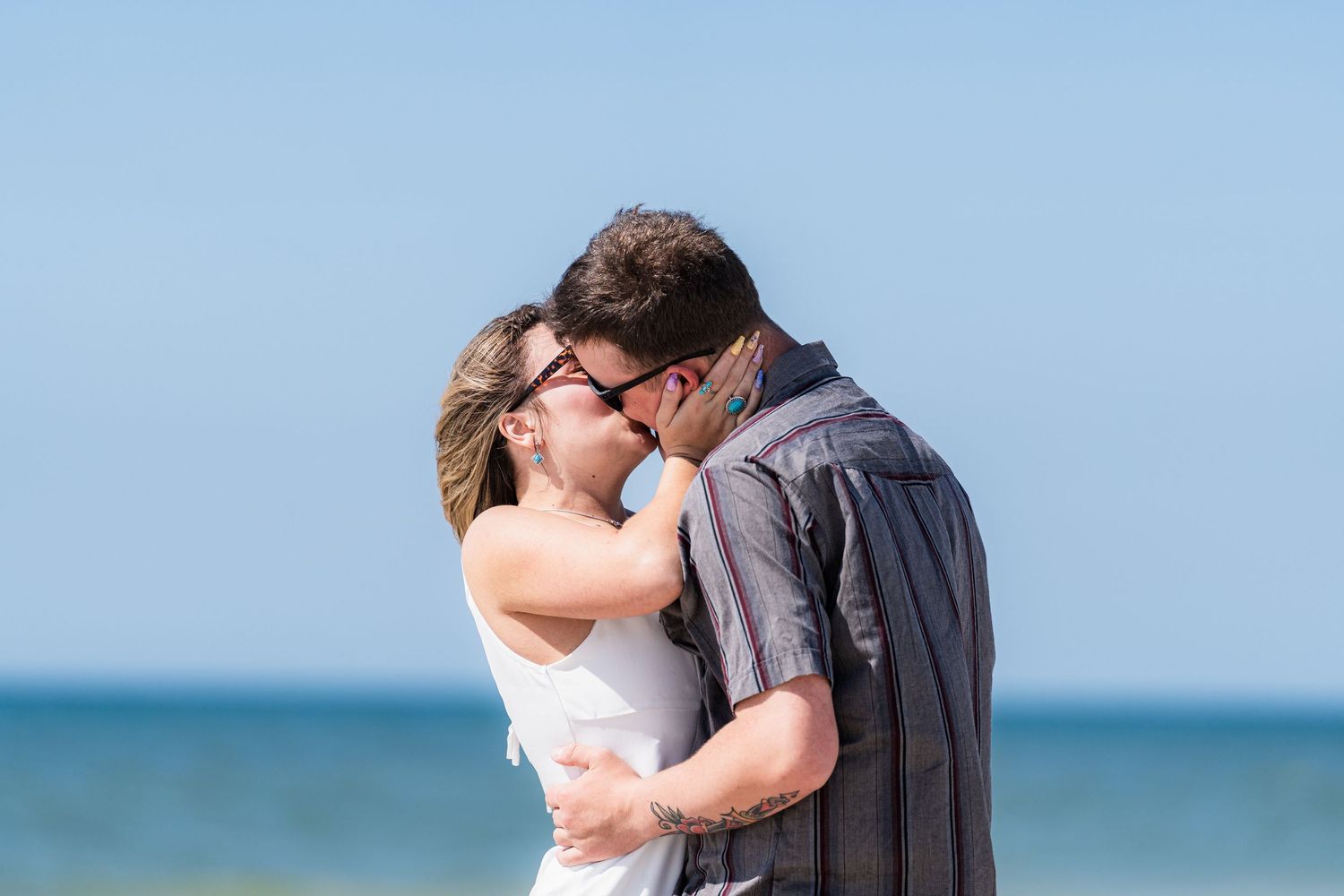 Couple sharing a romantic first kiss after beach engagement with ocean backdrop on a clear day.