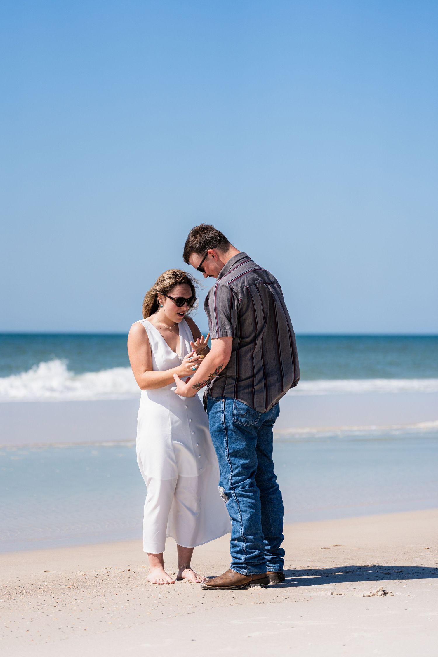 Couple standing on a sunny beach, woman in white dress and man in jeans, looking down together near ocean waves.
