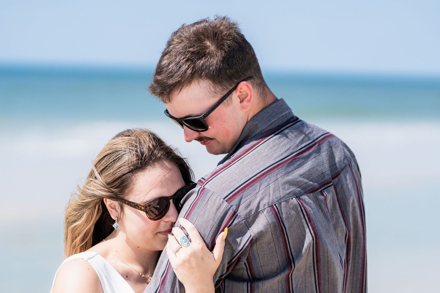 Romantic couple embracing on a beach, both wearing sunglasses on a bright sunny day.