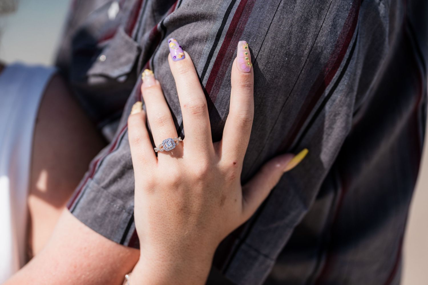 Close-up of a woman's hand with purple floral nails wearing an elegant engagement ring resting on a man's shirt.