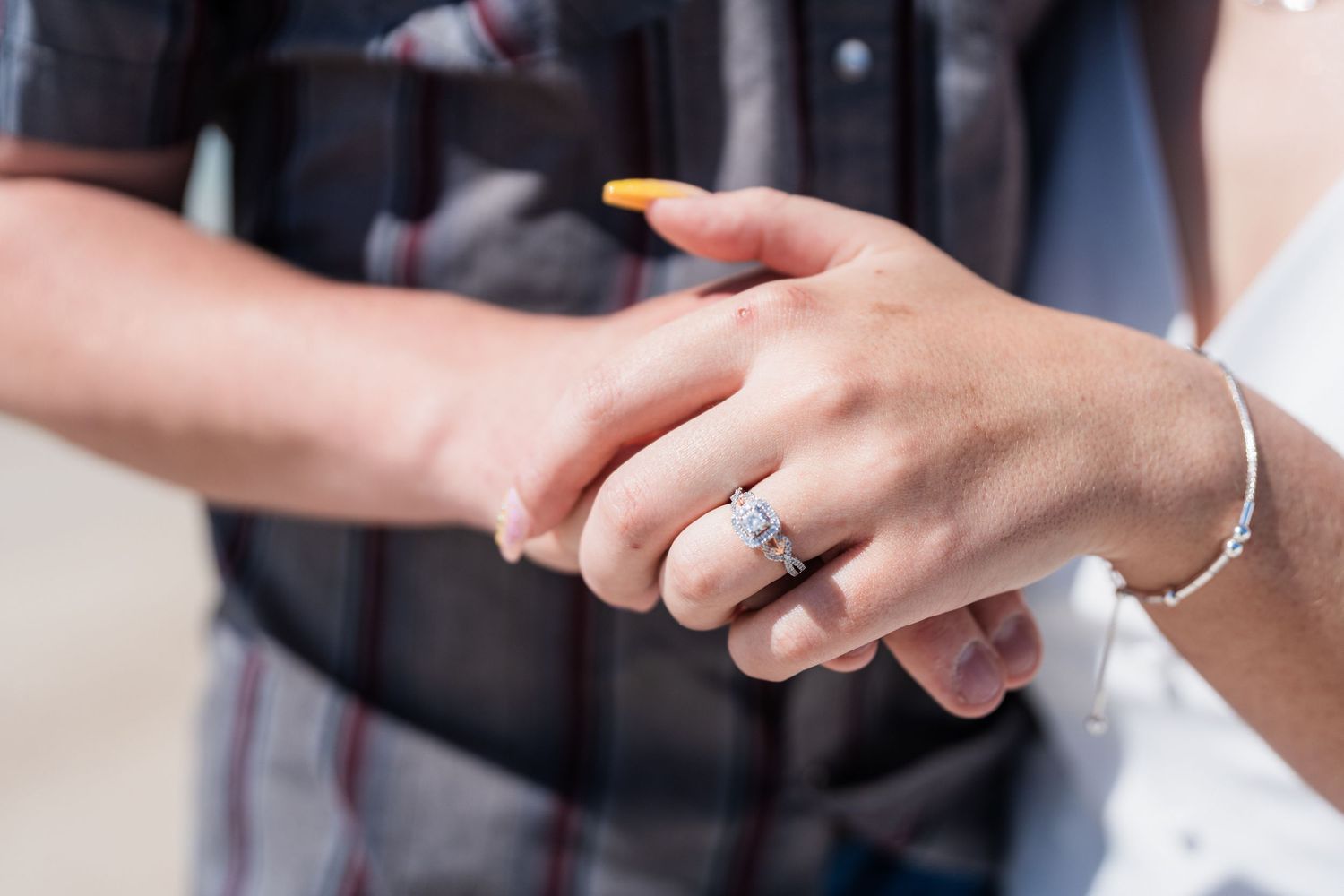 Close-up of engagement ring on woman's hand held by man in plaid shirt on beach.