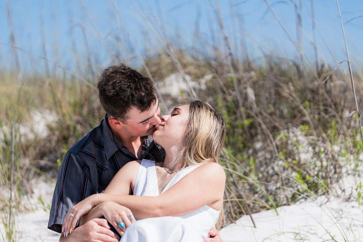 Couple sharing romantic kiss while sitting in white sand dunes surrounded by beach grass.