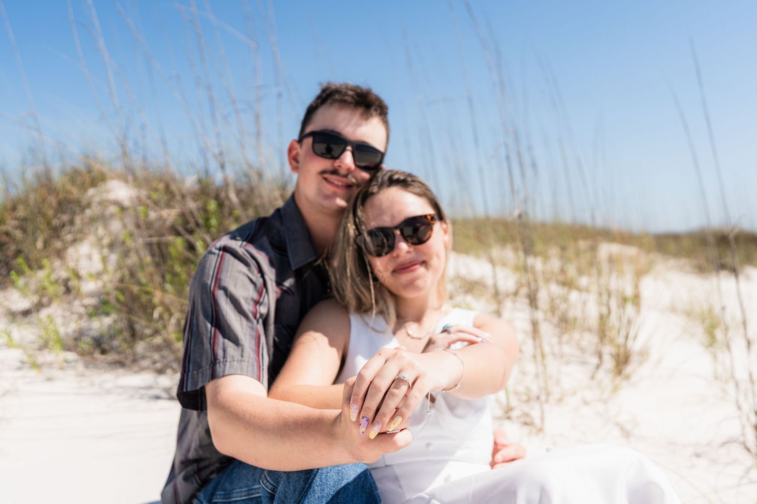 Couple in sunglasses embracing on a sandy beach with sea oats, enjoying a sunny day together.