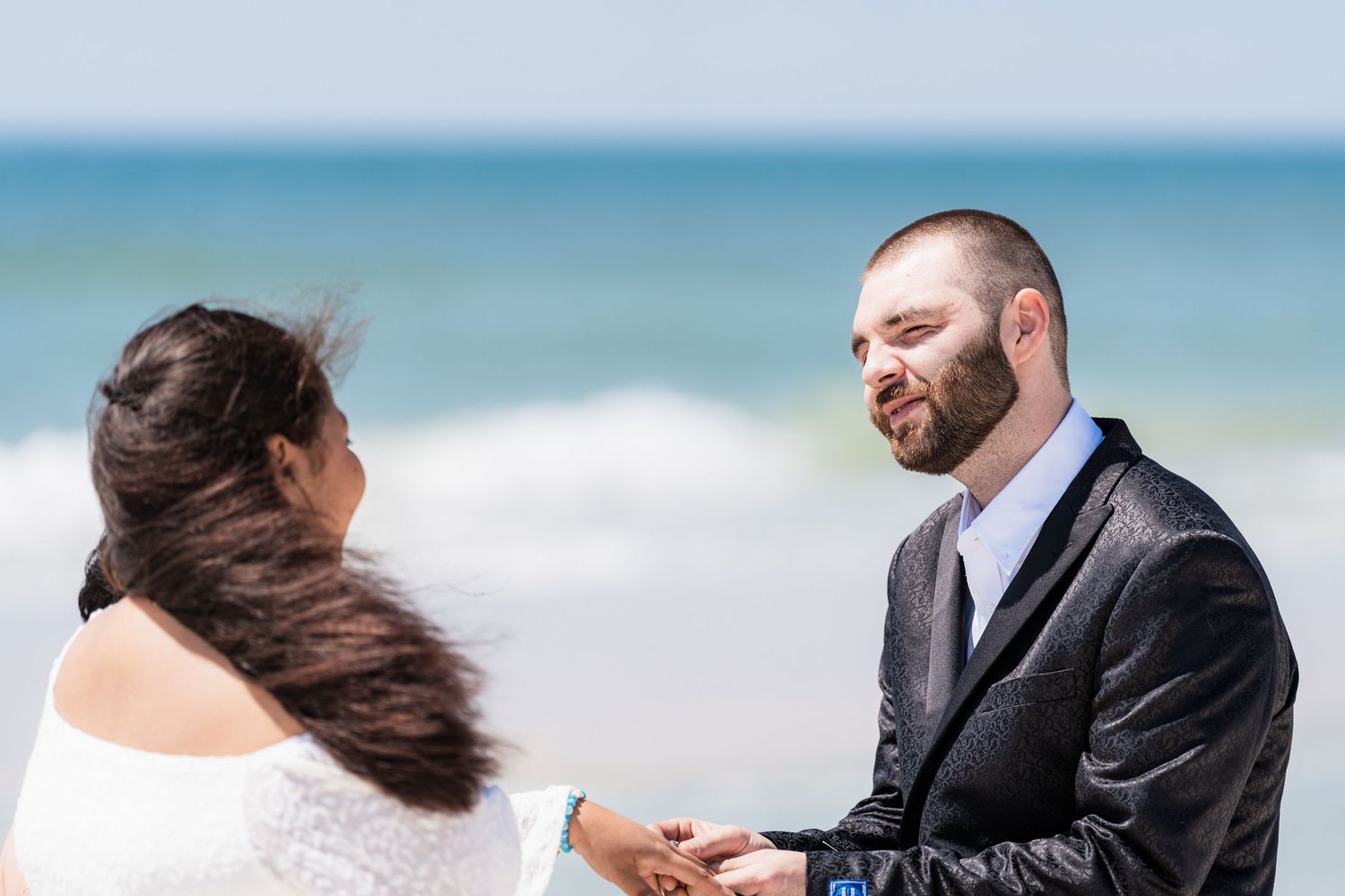 Groom smiling at bride during romantic beach wedding ceremony with ocean waves in the background.