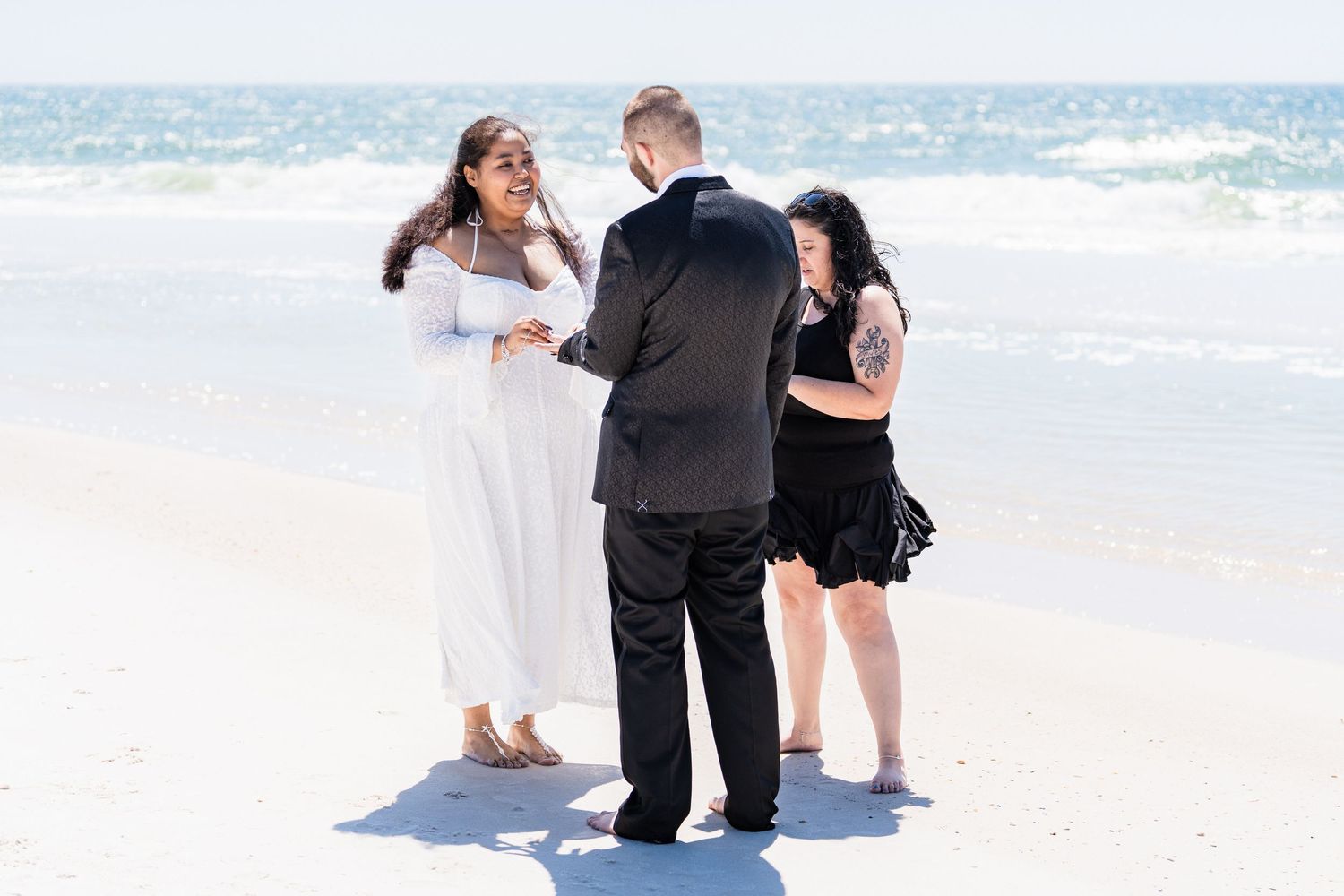 Couple exchanges vows during intimate beach wedding ceremony with officiant on white sand shore.