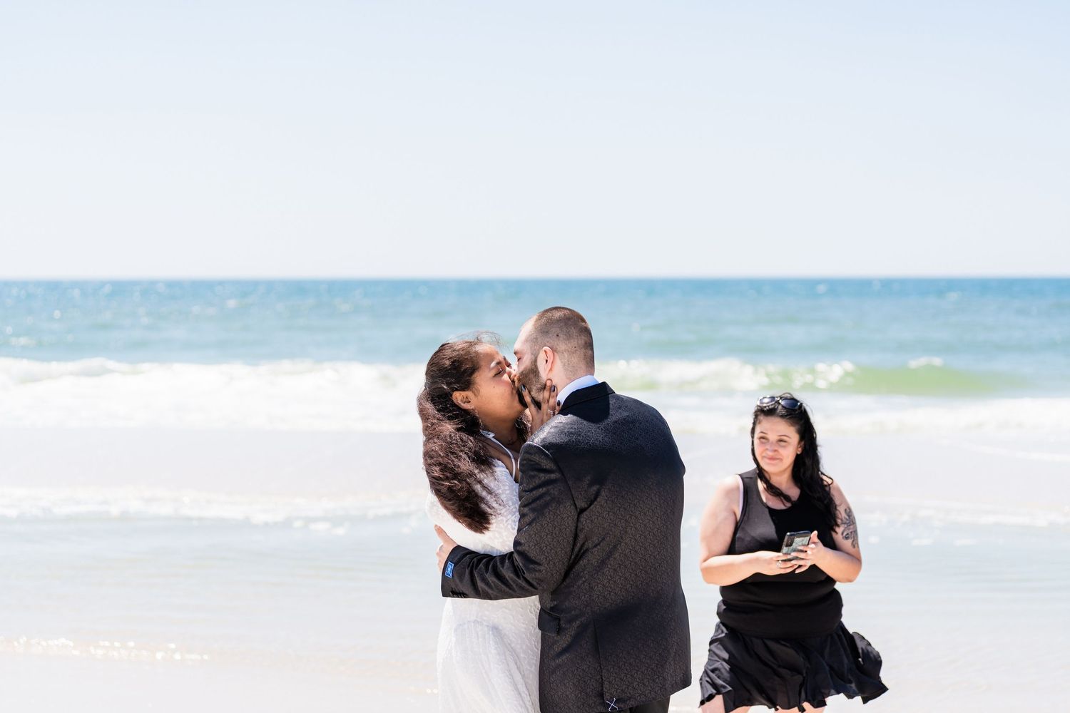 Couple sharing first kiss at beach wedding ceremony with officiant in black dress, ocean waves in background.