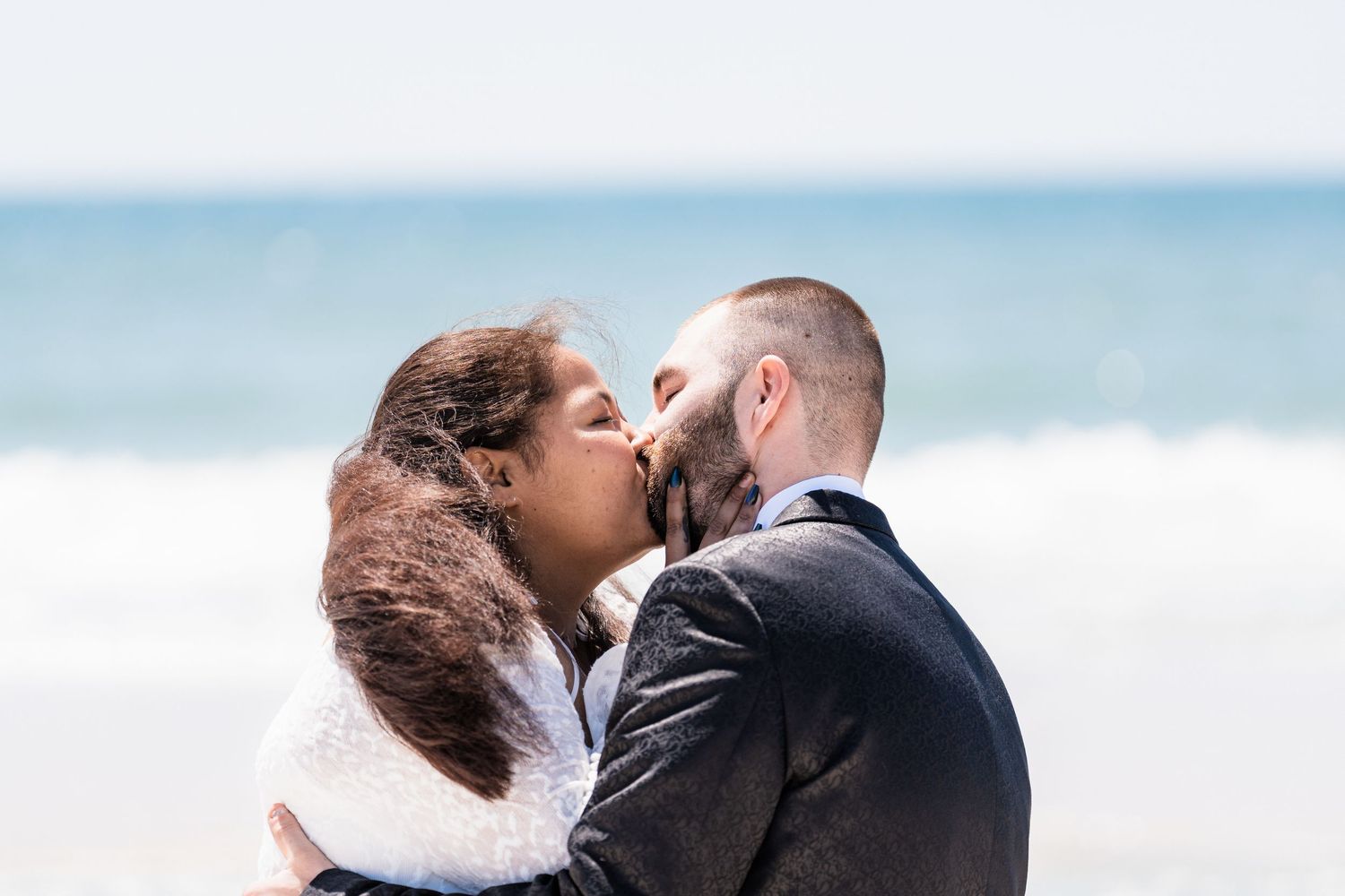 Newlyweds share first kiss as married couple on a breezy beach after outdoor wedding ceremony.