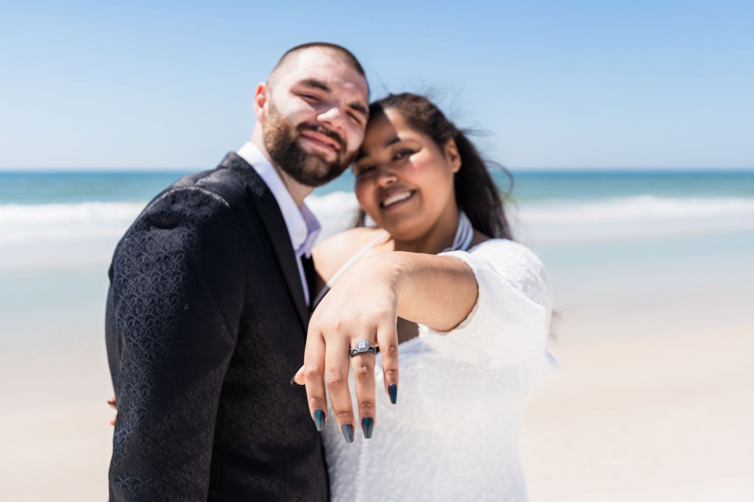 Happy couple on beach showing off engagement ring, smiling joyfully in wedding attire with ocean in background.