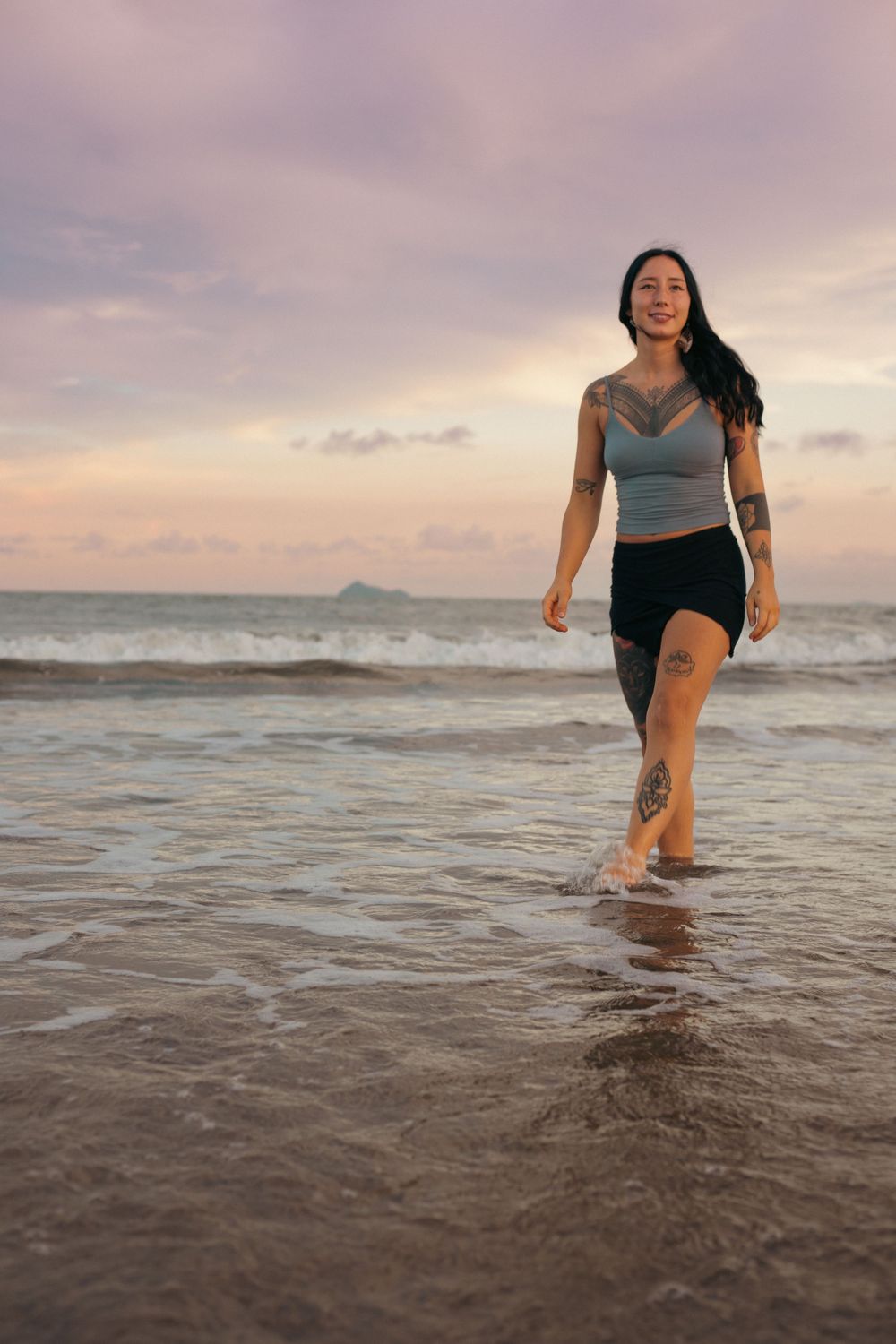 Tattooed woman in crop top and skirt walking barefoot on a beach at sunset with ocean waves.