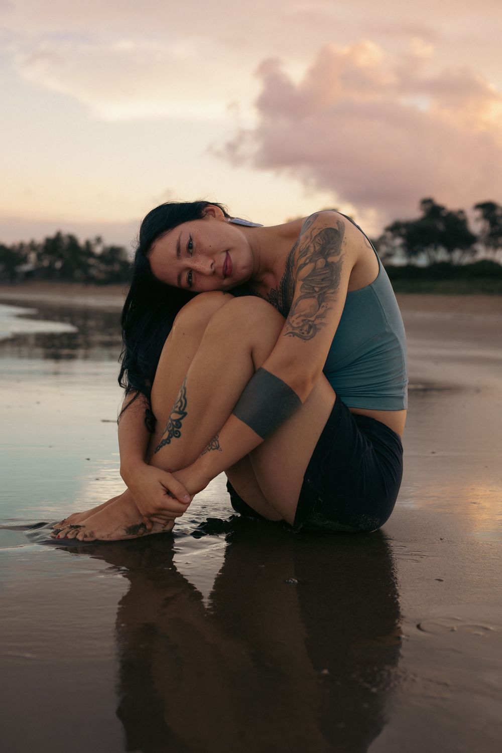 Tattooed woman sitting on wet beach sand at sunset, hugging her knees in a blue crop top and black shorts.