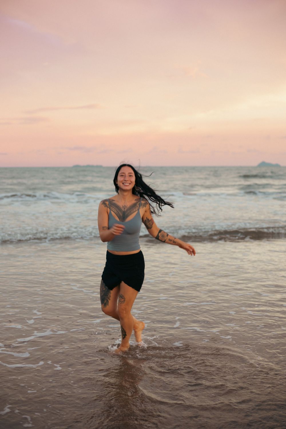 Smiling tattooed woman running in ocean waves at sunset beach.