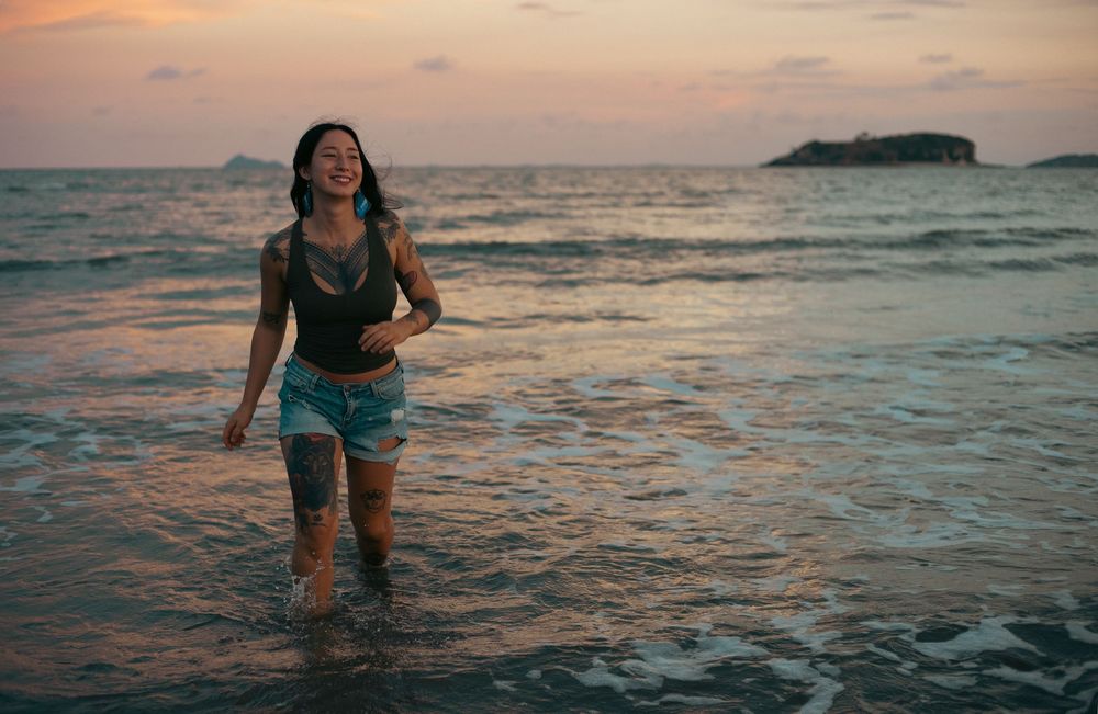 A smiling woman with tattoos wades through ocean waves at sunset, wearing denim shorts, with an island visible in the background.