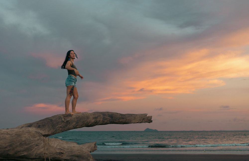 Woman standing on driftwood at beach during colorful sunset with dramatic orange and pink sky over ocean.